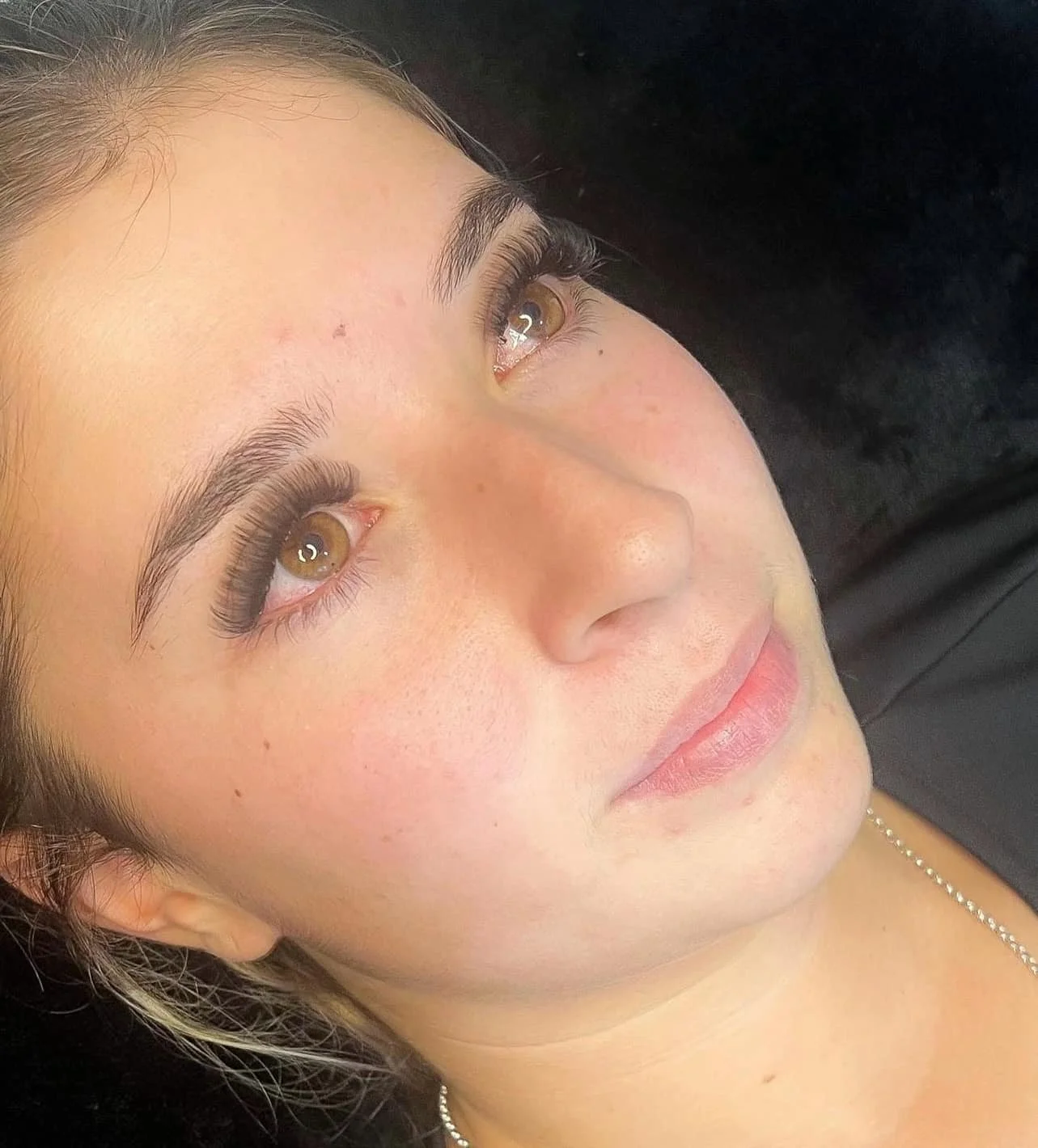 Close-up of a woman's face with brown eyes, dark eyebrows, and long eyelashes, lying on a black surface.