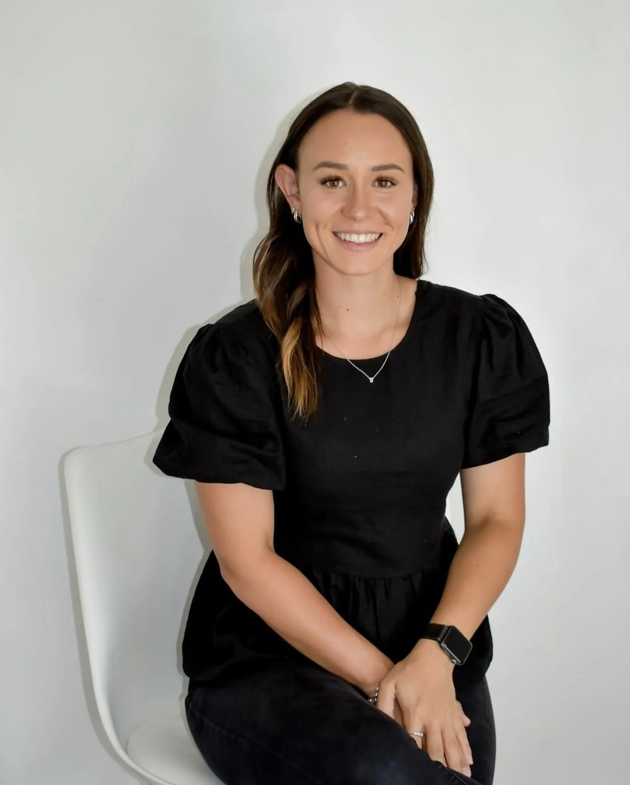 A woman with brown hair styled in loose waves, wearing a black puff-sleeve dress, seated and smiling in front of a plain white wall.