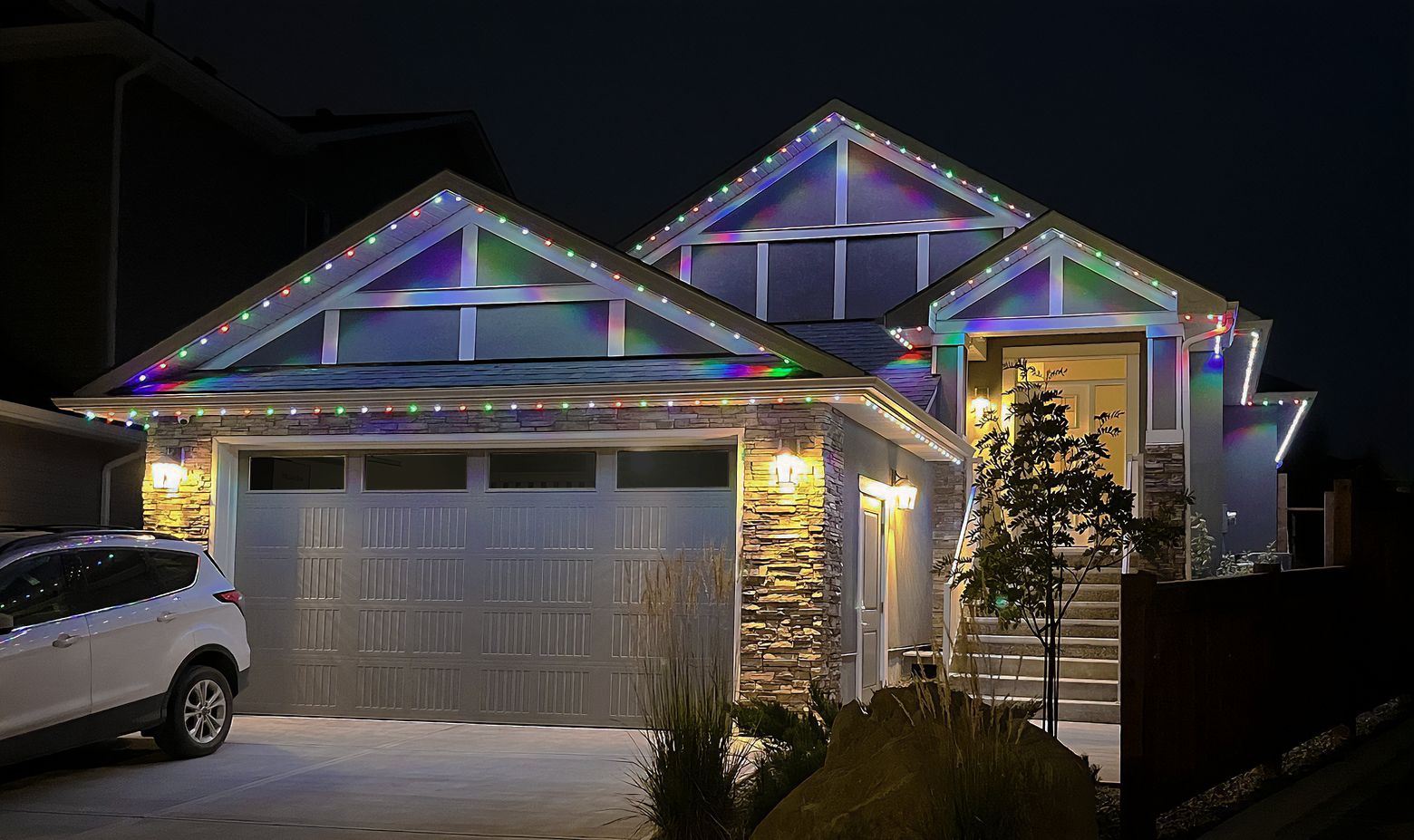 Night view of a modern house decorated with colorful Christmas lights on the roof and eaves. The house has a garage with a white security gate, a parked white SUV, and outdoor lighting near the front door. There are stairs leading to the front entrance, a small tree, and landscaping in the yard.
