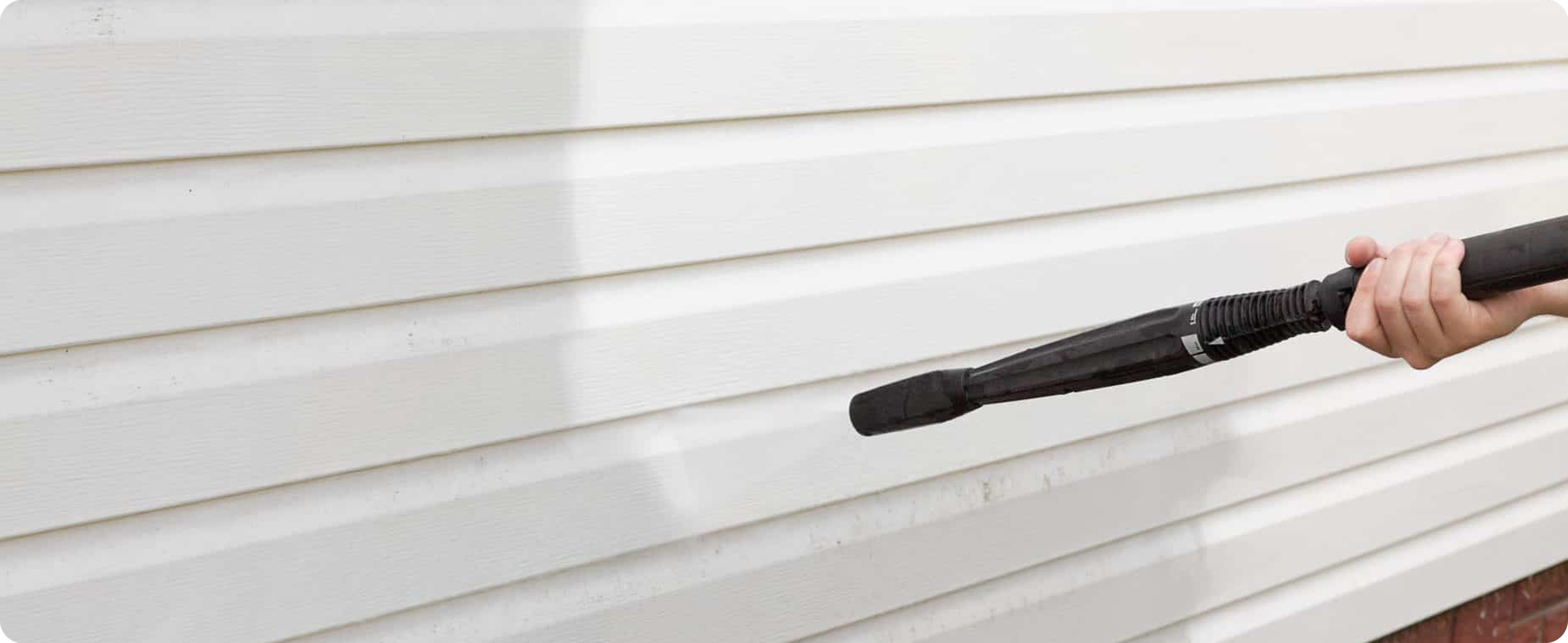 A close-up shot of a hand aiming a black pressure washer extension nozzle directly at dirty white vinyl siding to wash it down.