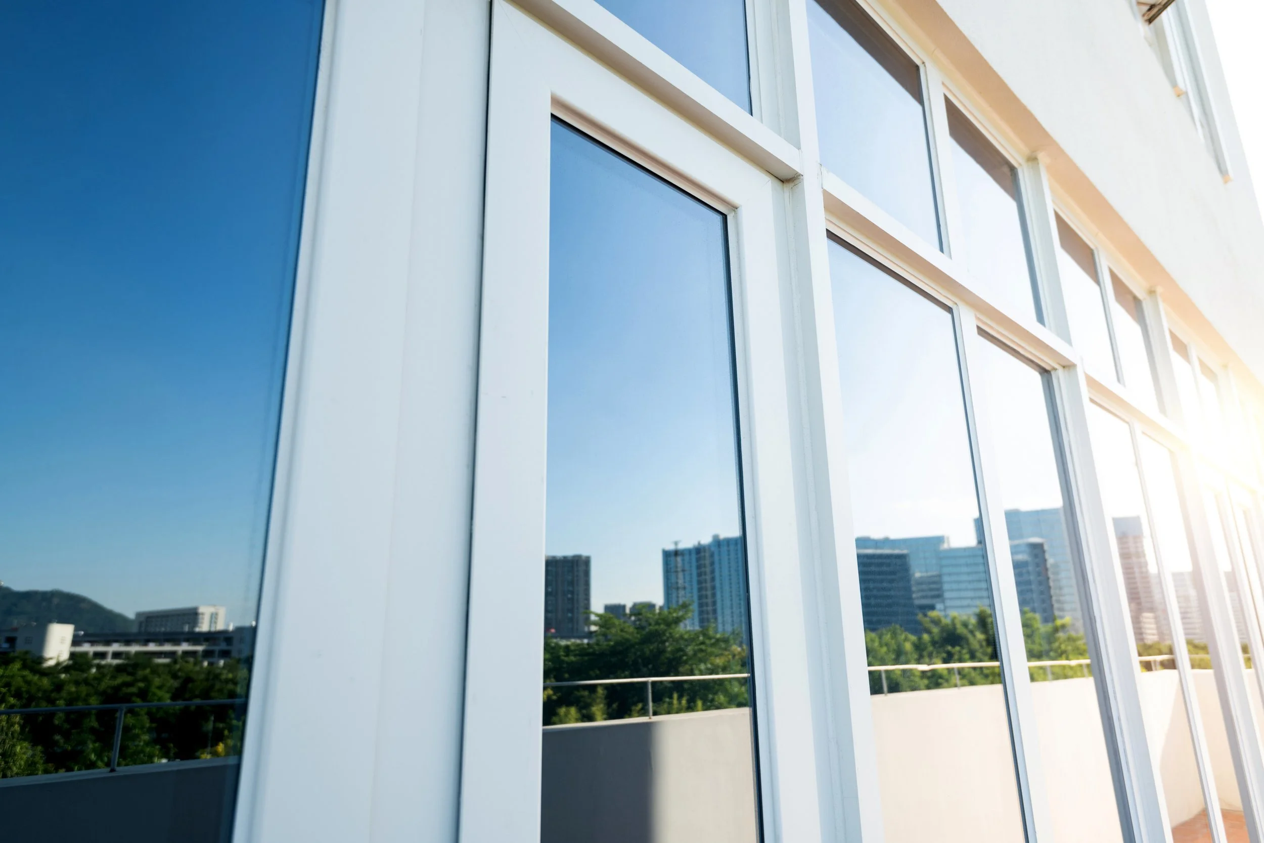 View of a modern building's glass windows reflecting an urban cityscape with trees and high-rise buildings, under bright sunlight.