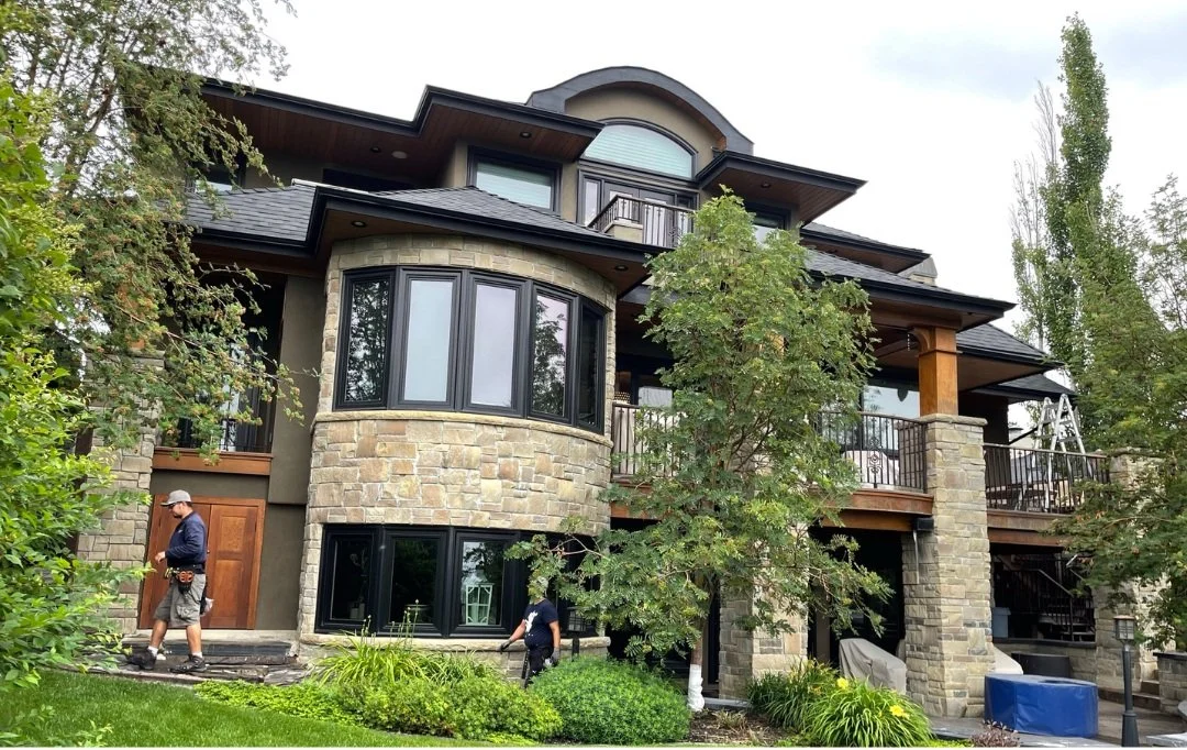 Large multi-story house with stone and wooden exterior, multiple balconies, and large windows surrounded by green trees and lawn.