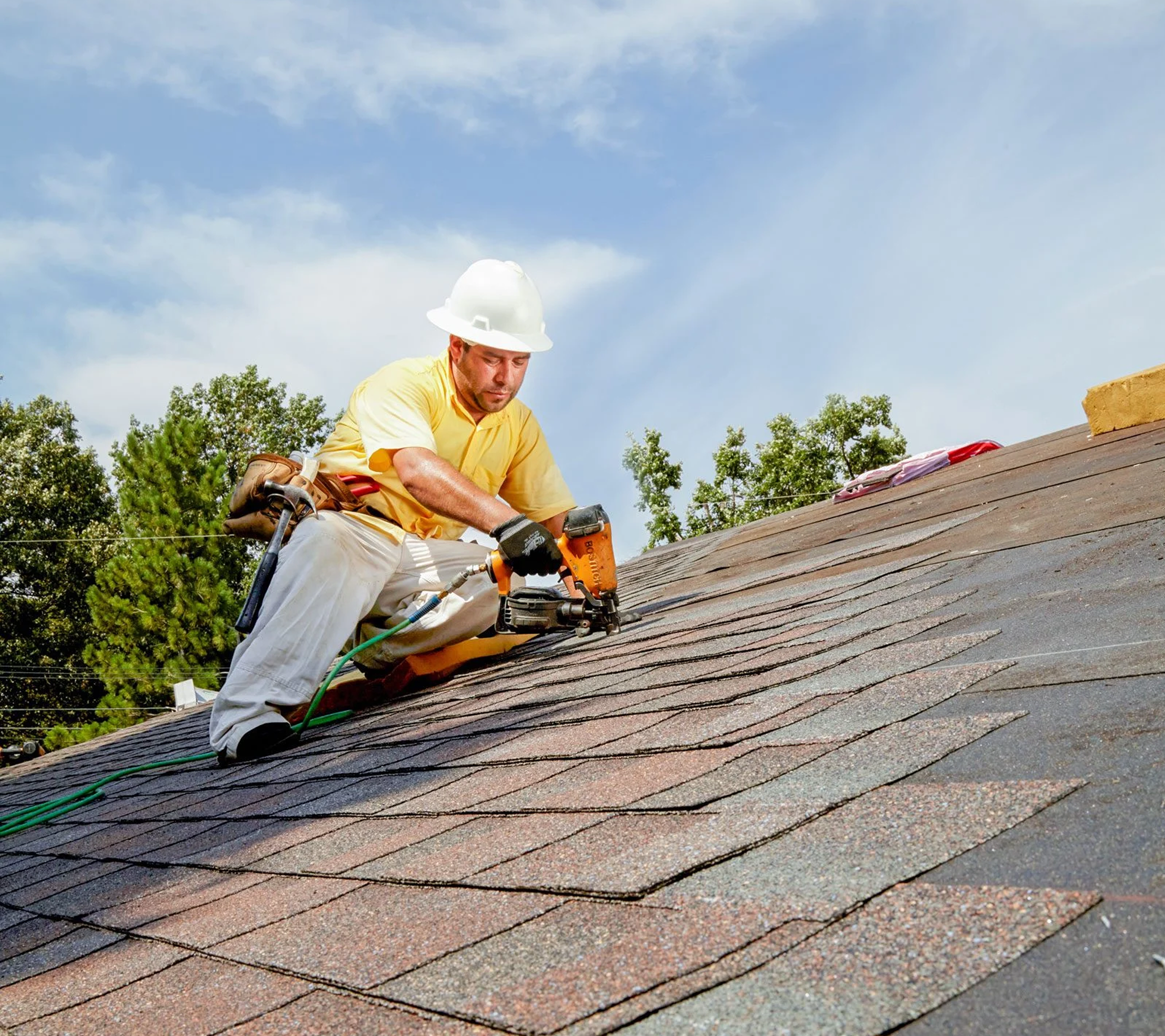 A man wearing a yellow shirt, white hard hat, and gloves using a power tool to work on a sloped roof during daytime. The roof is covered with shingles, and the background shows trees and a partly cloudy sky.