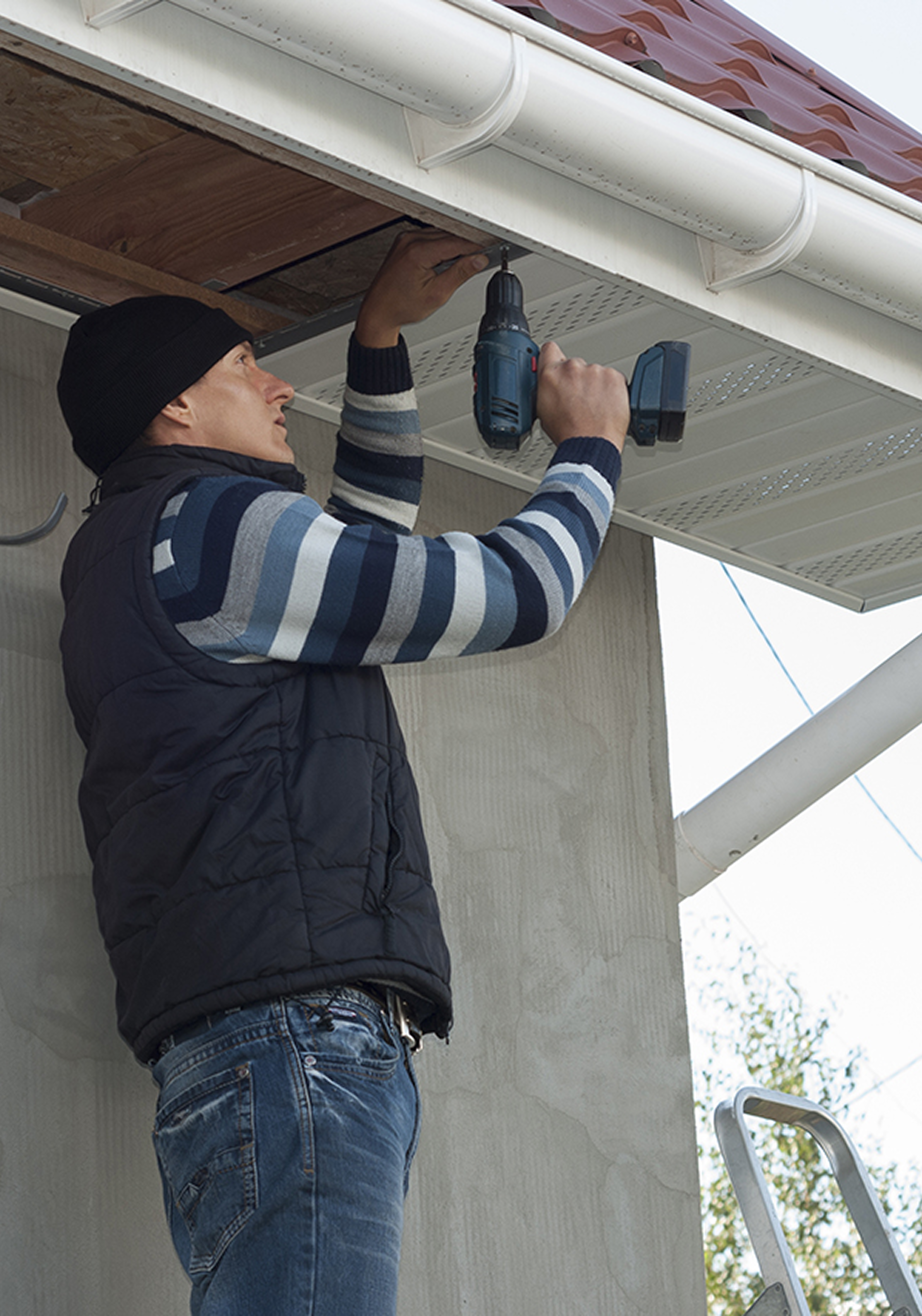 A man wearing a black beanie, striped sweater, and jeans is standing on a ladder while using a cordless drill to work on the underside of a house's roof or soffit.