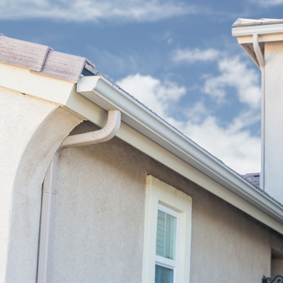 Close-up of a house's beige stucco wall, showing roof gutter and downspout, with a window and part of the roof visible under a partly cloudy sky.