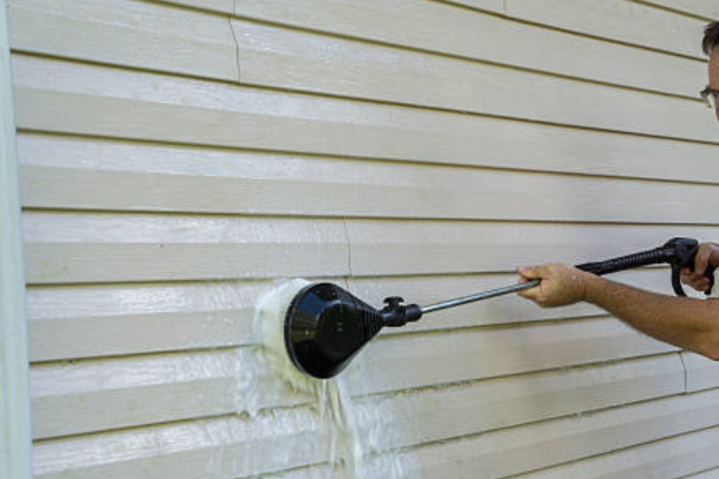 Person power washing beige vinyl siding with a high-pressure cleaning tool.