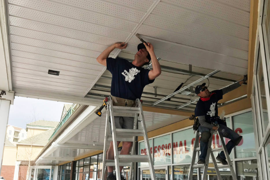 Two workers installing or repairing a suspended ceiling inside a storefront with large windows.