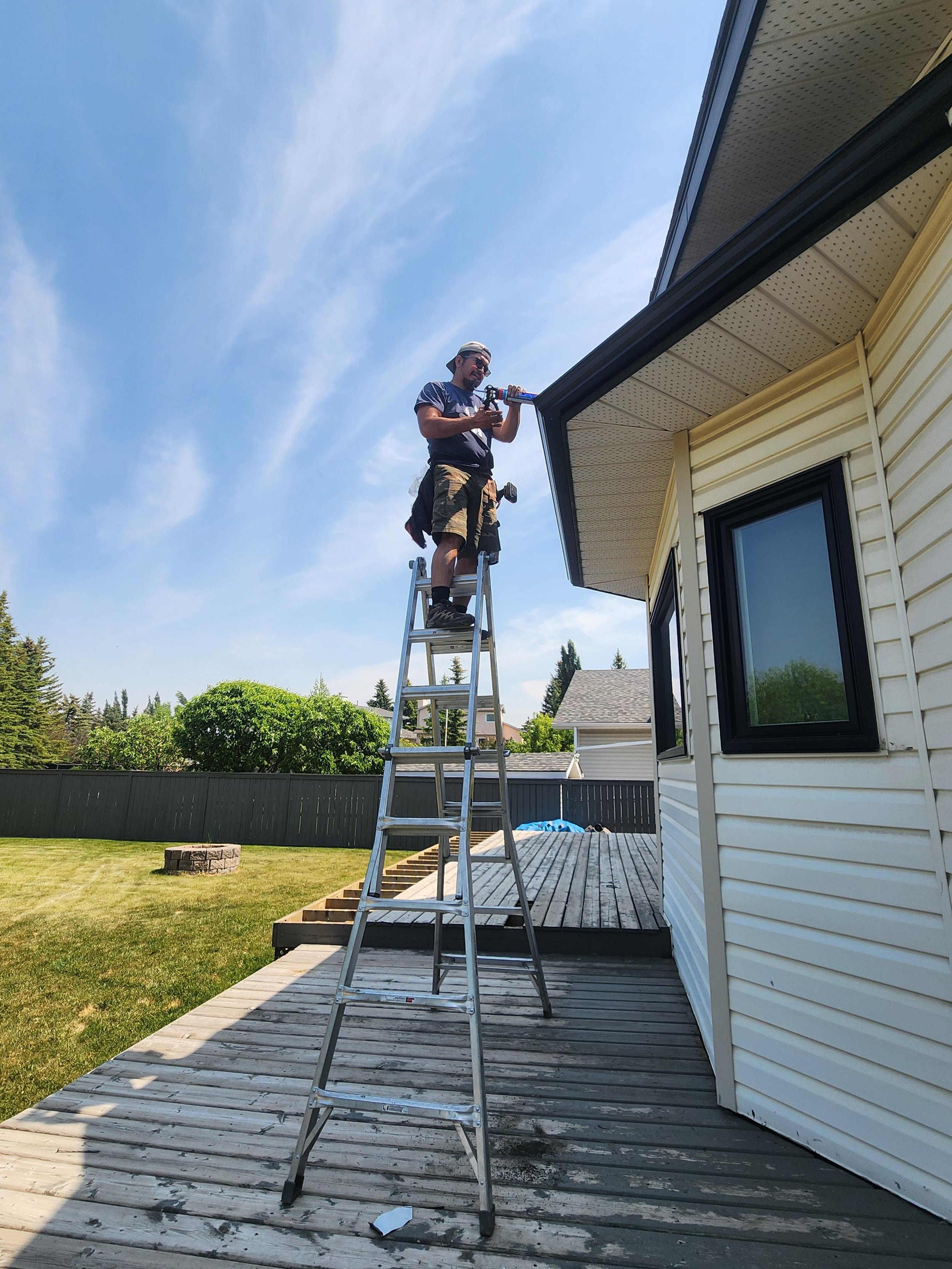 Man standing on a ladder repairing or installing a gutter on the side of a house on a sunny day.