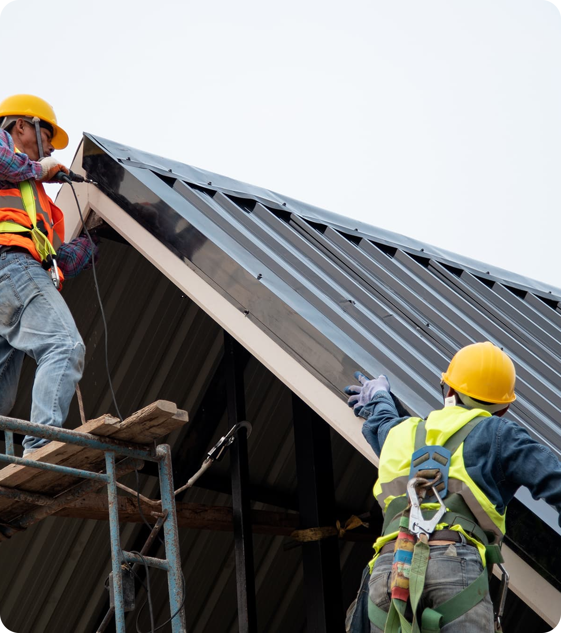 Two construction workers on scaffolding installing solar panels on the roof of a building. One is wearing a yellow helmet, a high-visibility vest, and safety gloves. The other worker is wearing a yellow helmet, a high-visibility vest, and has tools hanging from a belt.