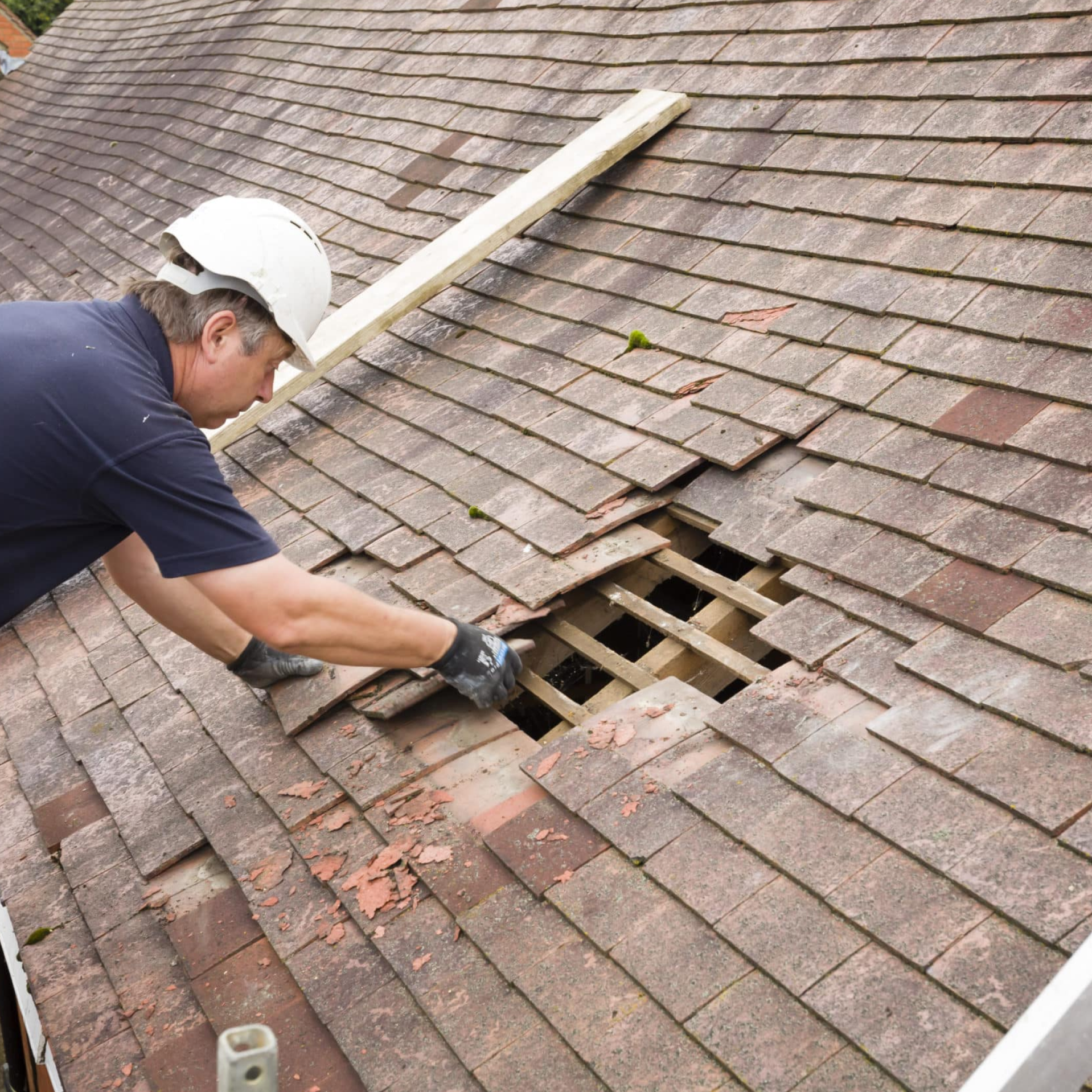 A person wearing a white safety helmet and dark clothing bent over on a sloped brick roof during repair, with a wooden support beam placed across the roof to assist in the roofing work.