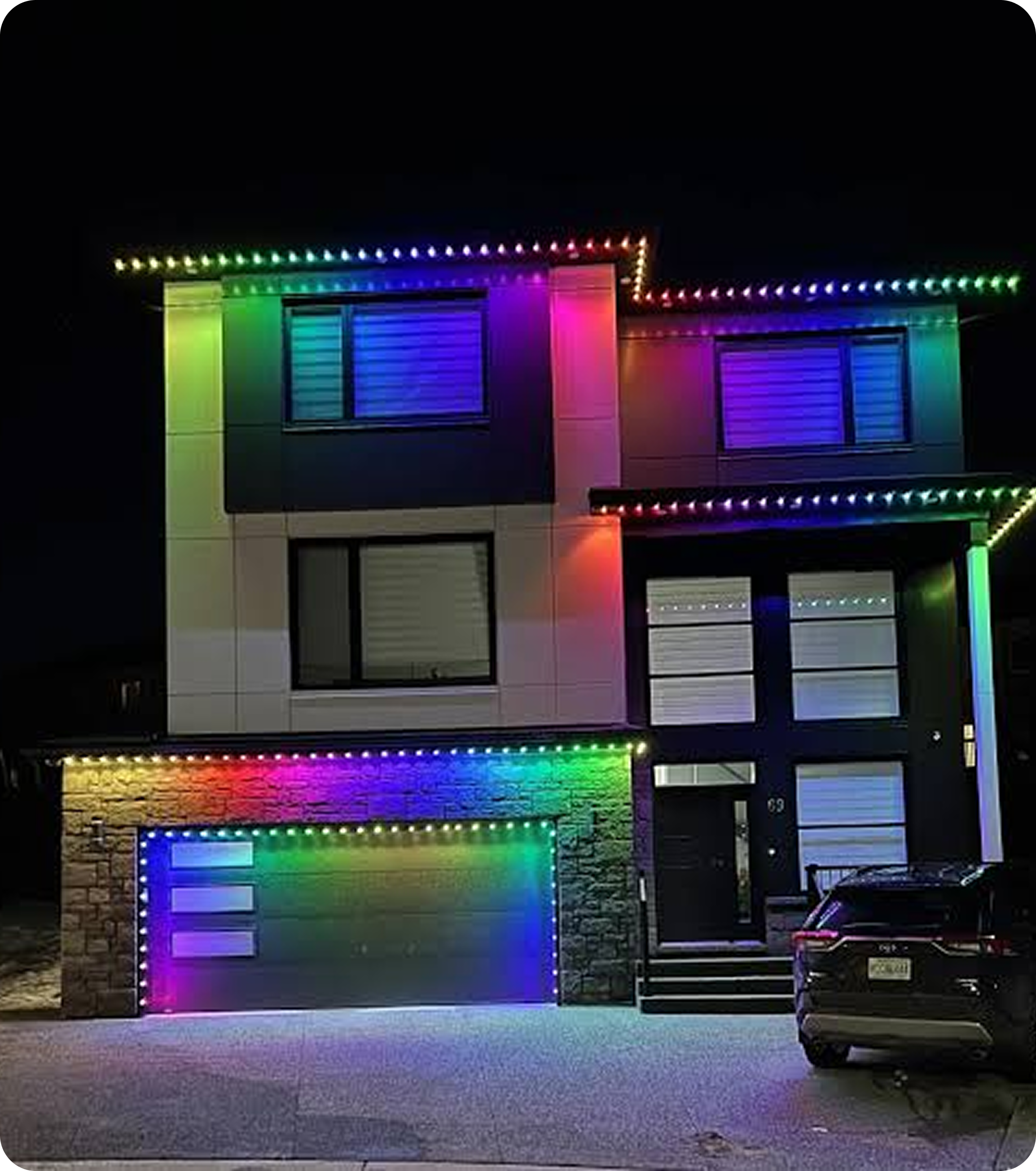 Nighttime view of a house with two large windows, decorated with colorful LED Christmas lights outlining the roof and around the windows, creating a festive display. A parked car is visible on the right side of the image.