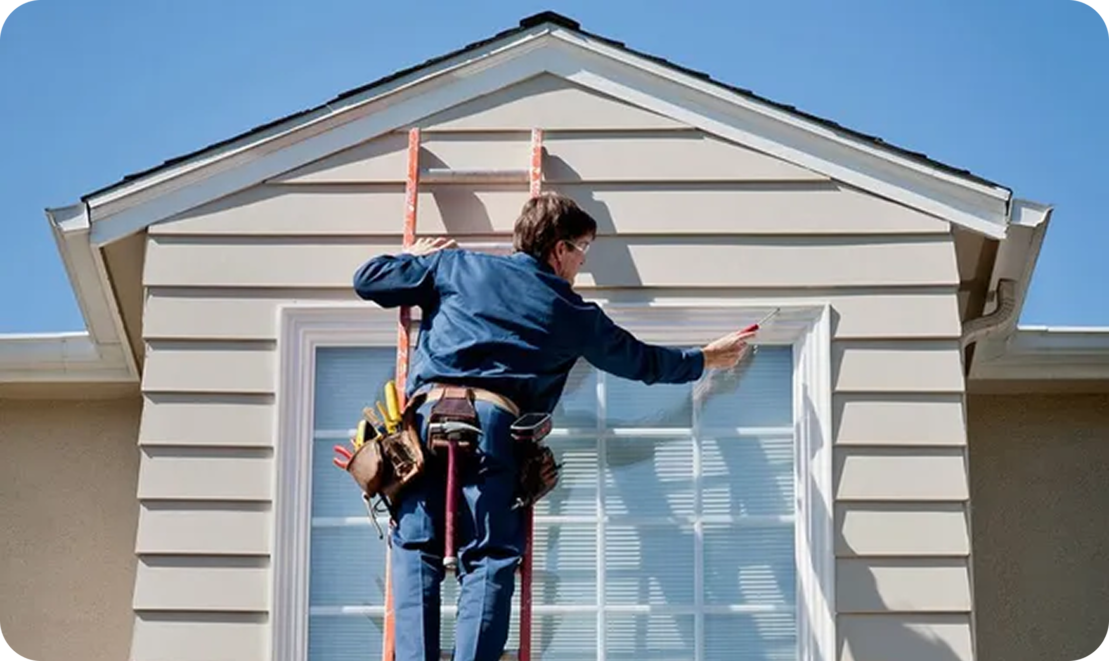 A house painter in a blue shirt painting window trim on a house, using a ladder and wearing a tool belt under a blue sky.