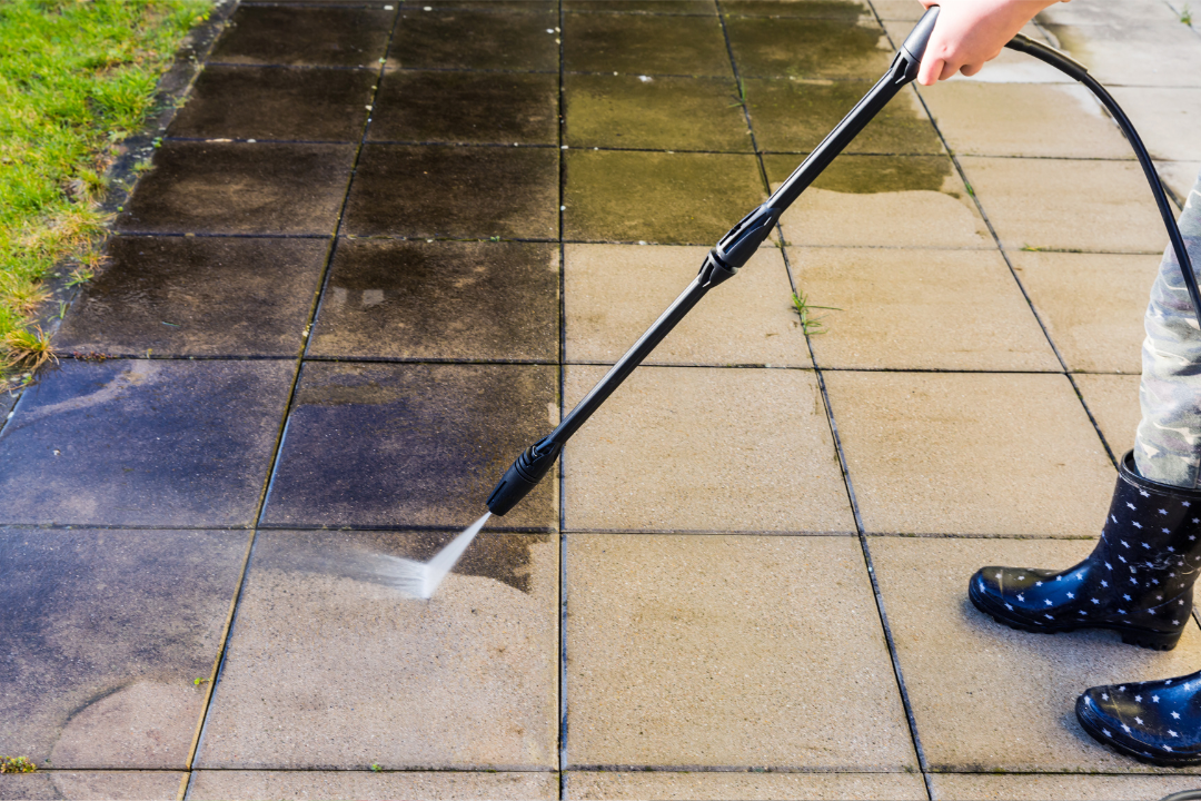 A person using a pressure washer to clean a paved sidewalk, with half of the sidewalk covered in dirt and grime and the other half clean.