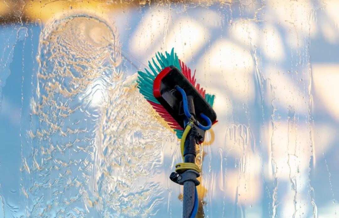 Window being cleaned with a scrub brush attached to a pole, water droplets on the glass, during daylight.