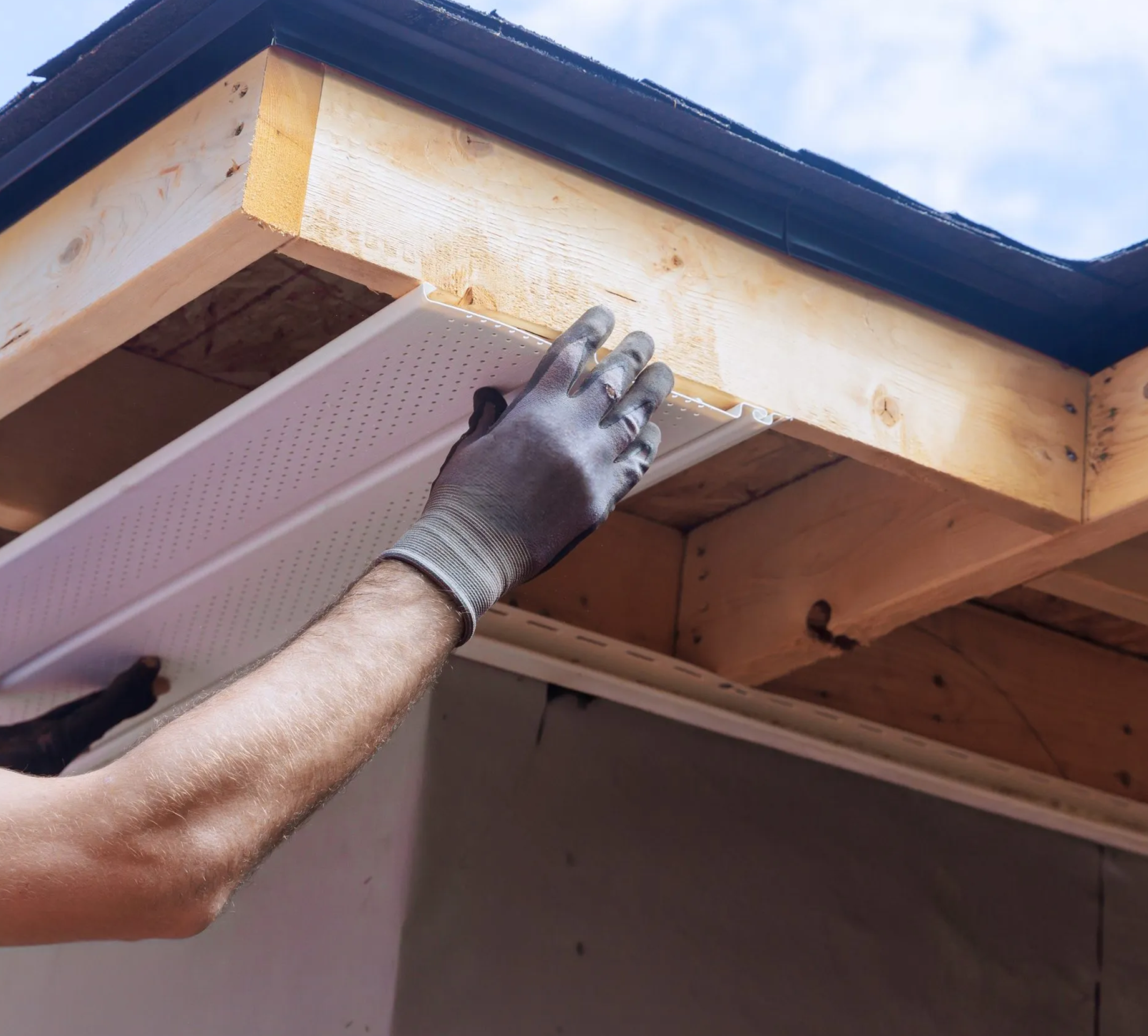 A person with a gray glove installing a beige perforated pegboard under a wooden shelf inside a construction site, with a black metal drip edge on the roof above.