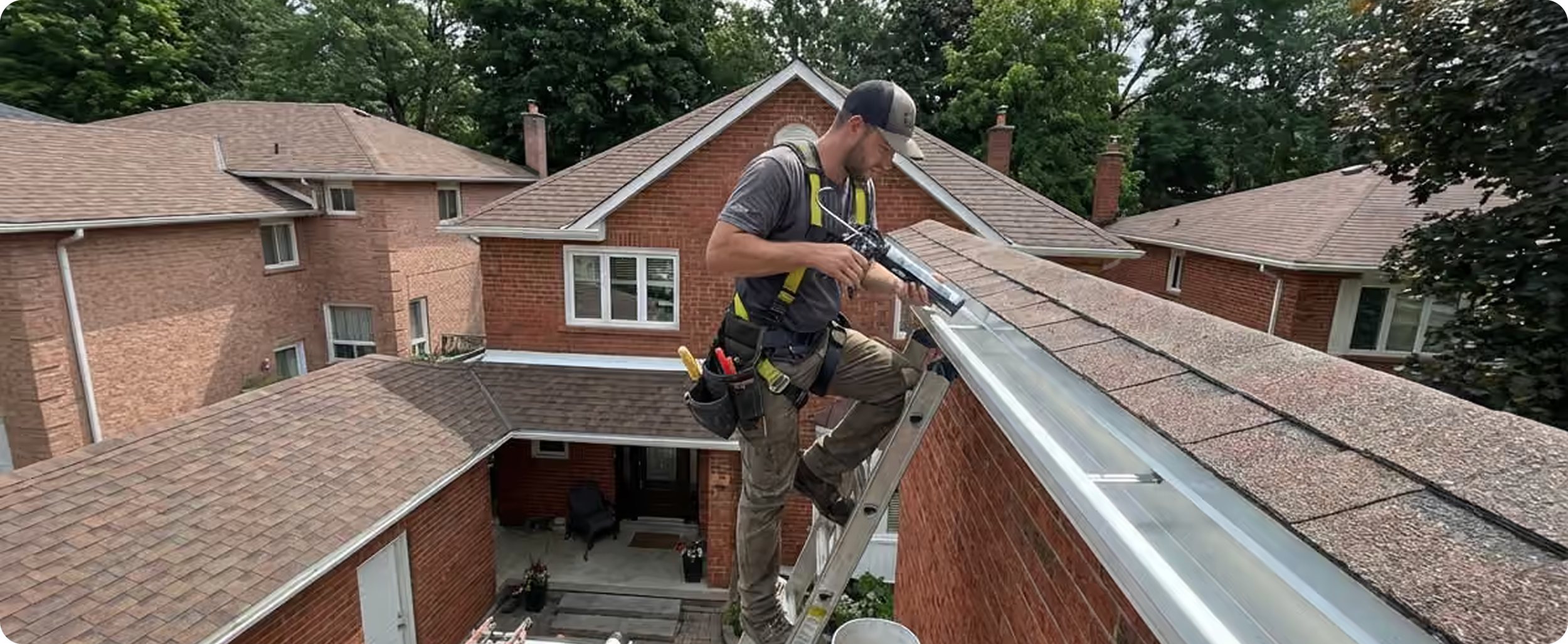 A man working on a roof, standing on a ladder, using a caulking gun or sealant applicator on the edge of a brick building with neighboring houses and trees in the background.
