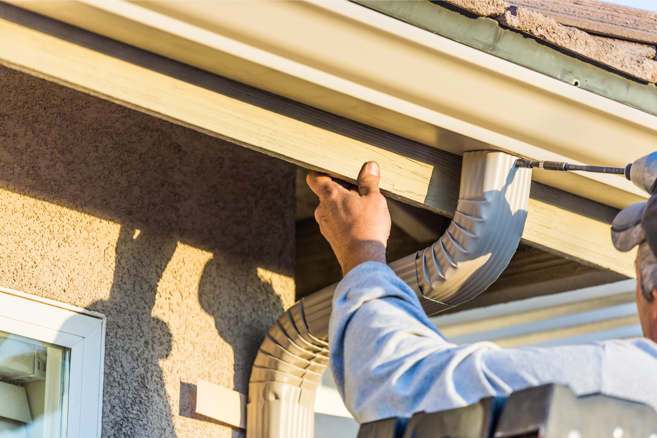 Person installing or inspecting a wooden board on the exterior of a house with a window, in sunlight.