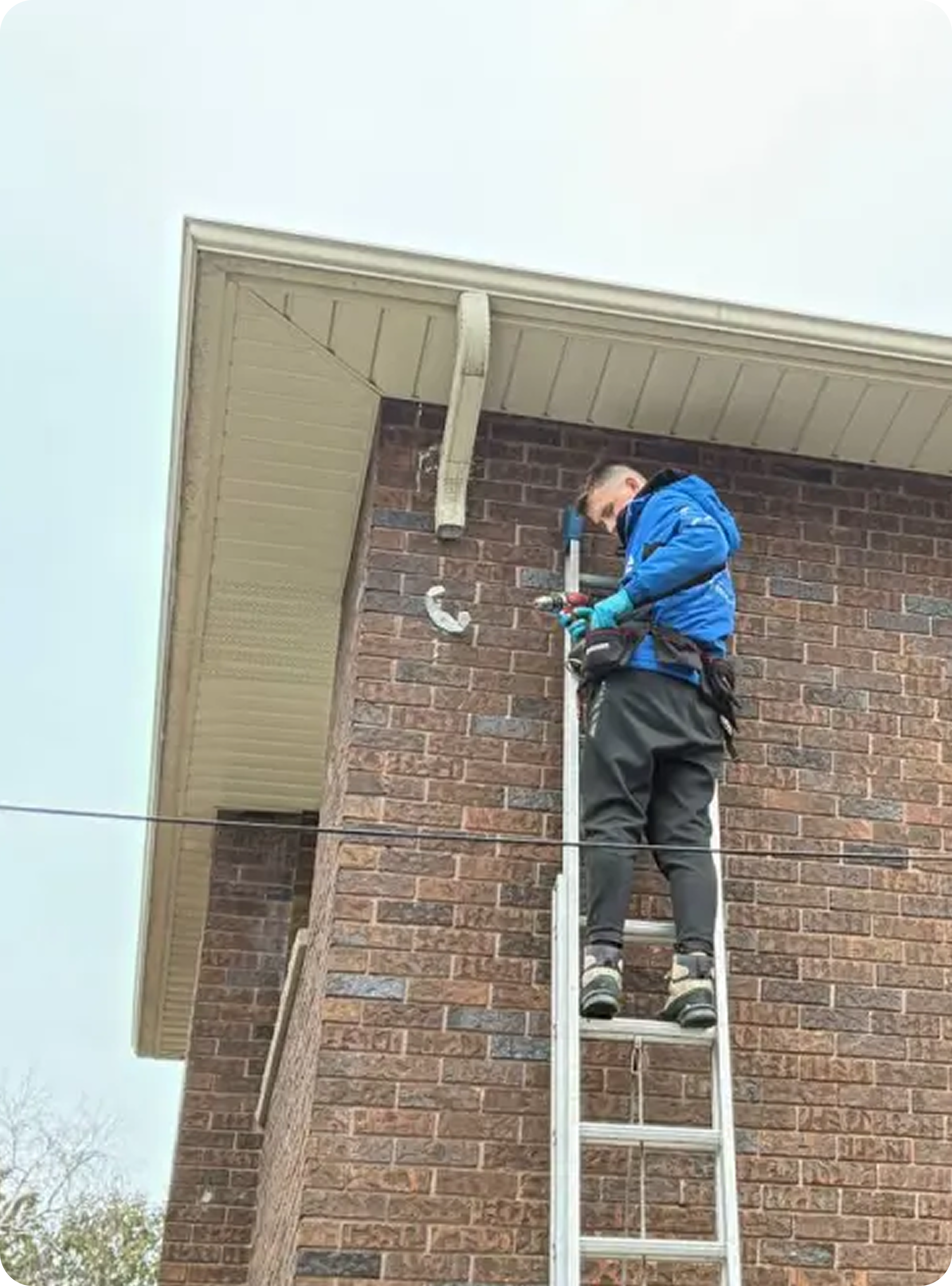 A person wearing a blue jacket and gloves standing on a ladder, working on the gutter of a brick house.