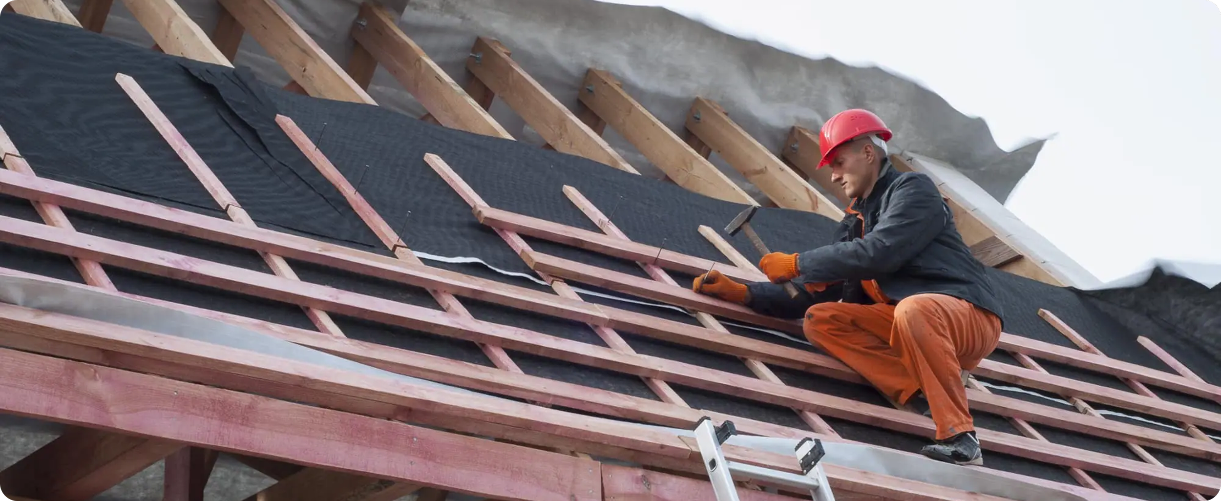 A construction worker wearing a red safety helmet and orange pants is kneeling on a roof structure, working on the wooden framework during roofing construction.