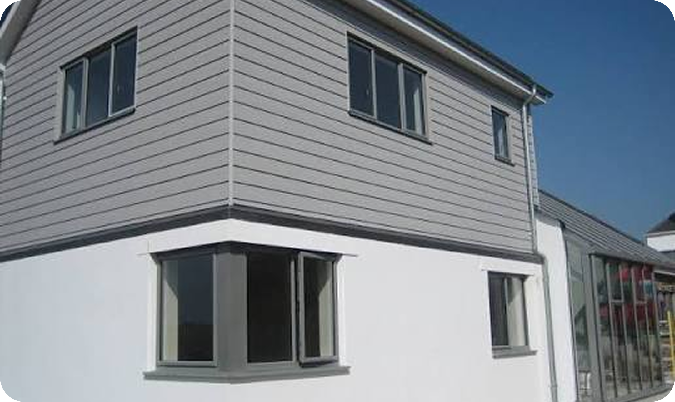 A modern, light-colored house with horizontal siding, several windows, and a distinctive bay window protruding from the lower level. The house has multiple sale signs on the windows, indicating it is for sale. The sky is clear and blue.