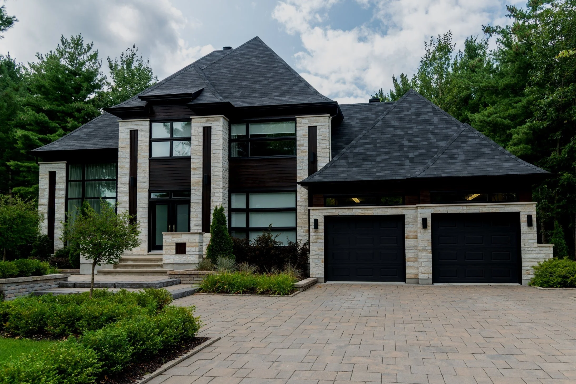 Modern two-story house with a dark roof, beige brick and dark wood exterior, large windows, attached garage, front steps, and landscaped yard with greenery and trees.