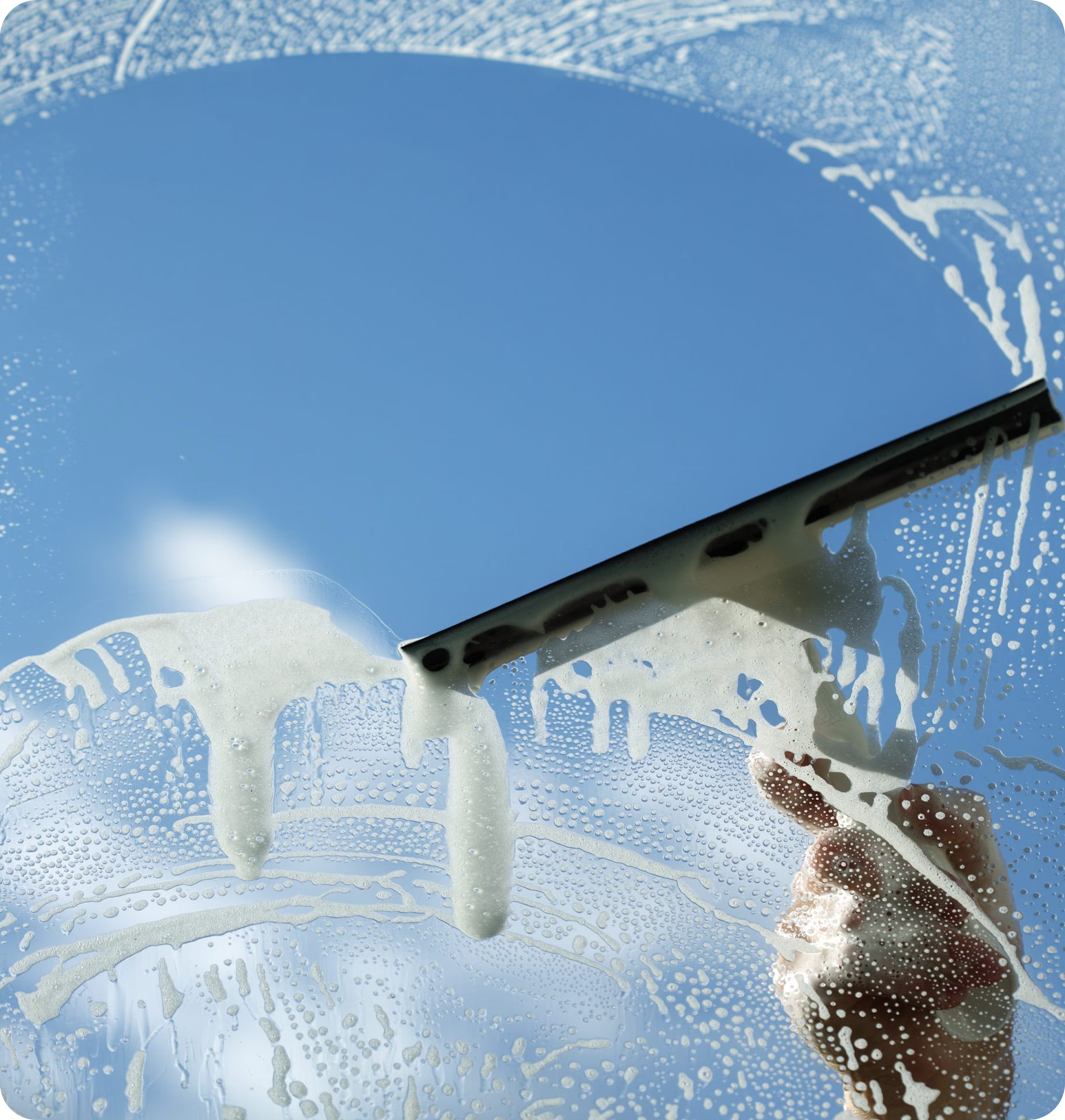 A close-up view of a hand holding a squeegee and wiping away soapy foam from a glass window pane, revealing a bright blue sky behind it.