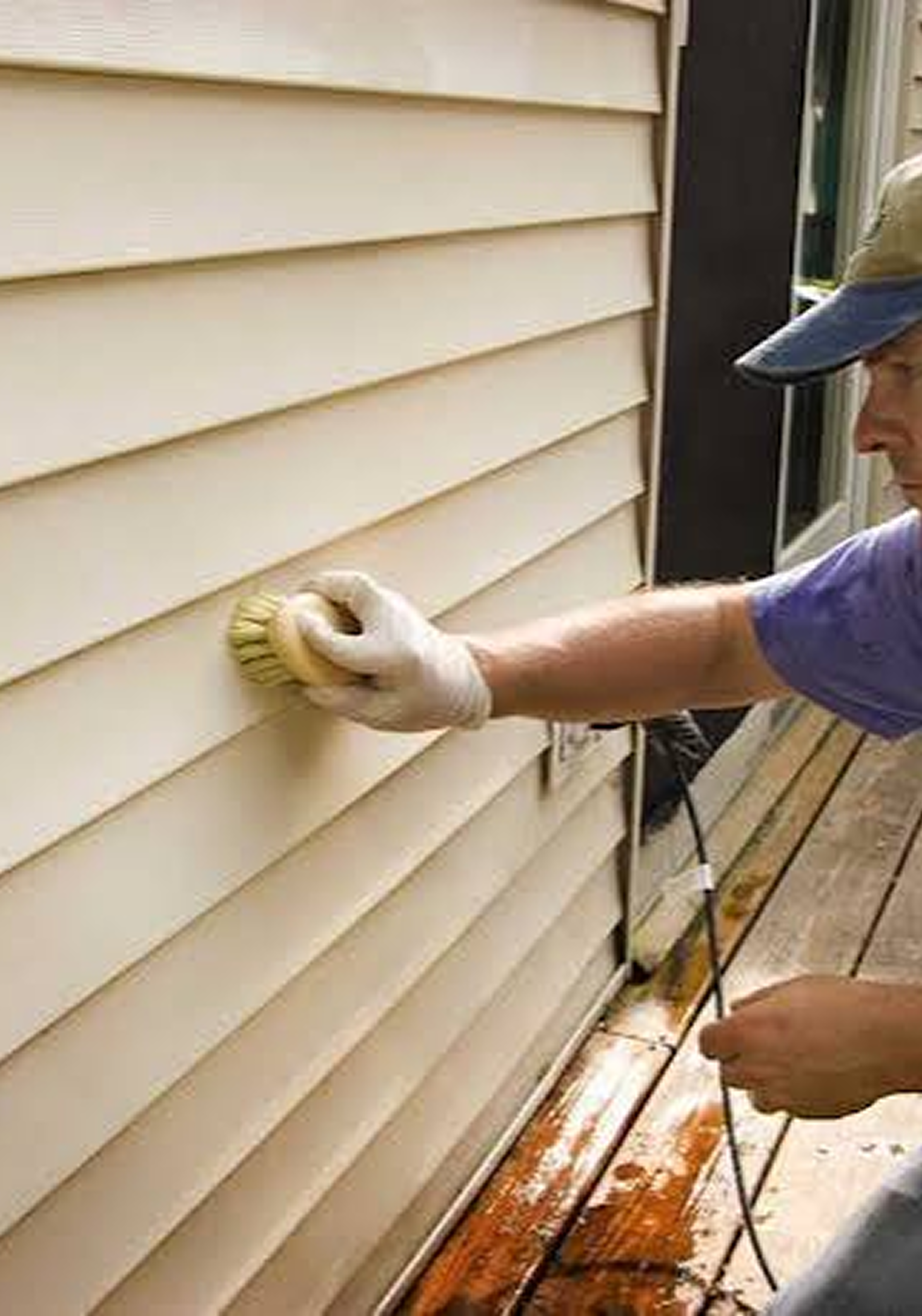 A person wearing a blue shirt, white gloves, and a baseball cap scrubbing the exterior beige siding of a house with a scrub brush, standing on a wooden deck.