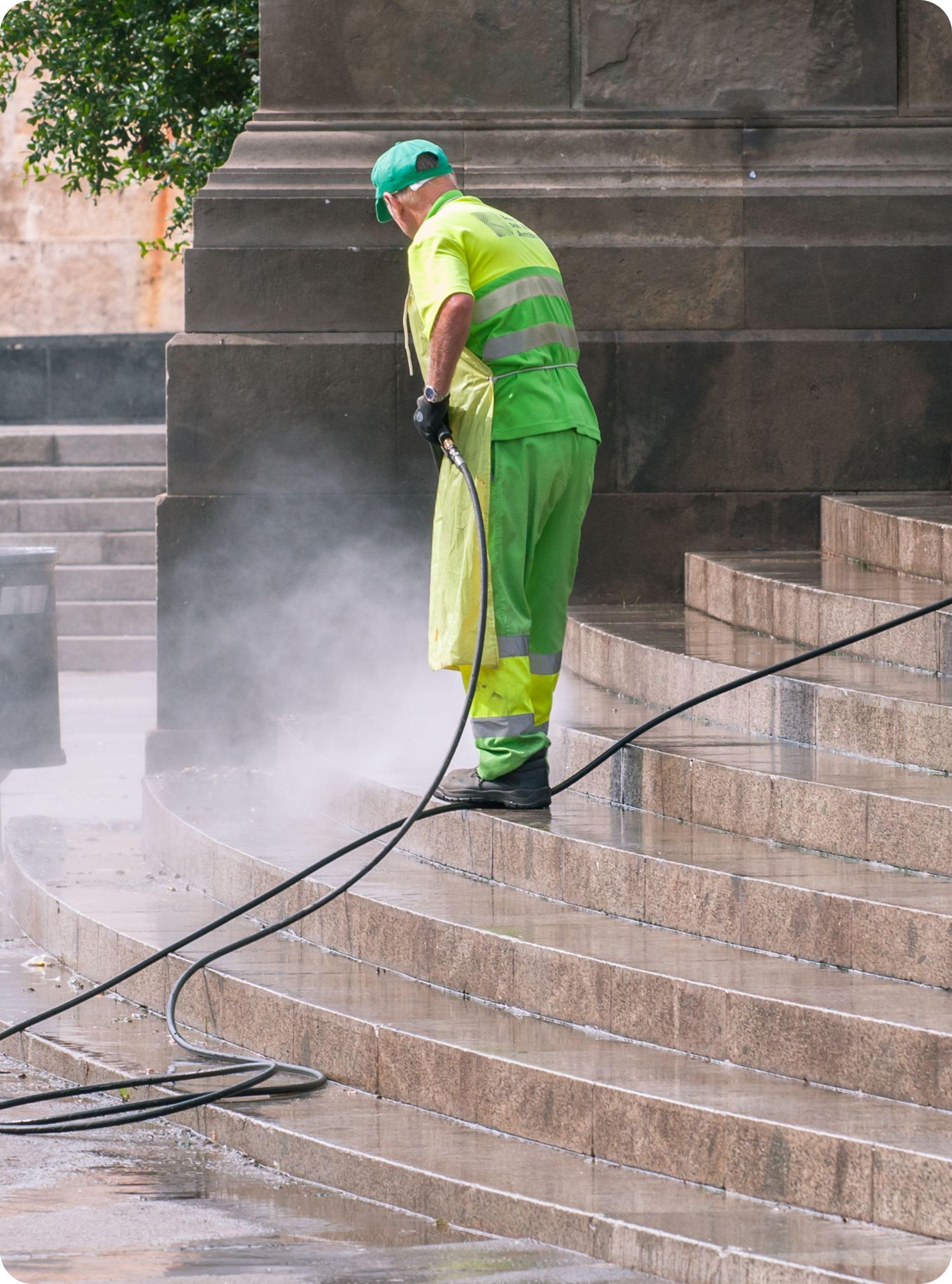 A worker wearing a high-visibility green uniform, cap, and apron standing on wide stone stairs, using a pressure washer hose that sprays a misty water jet onto the steps.