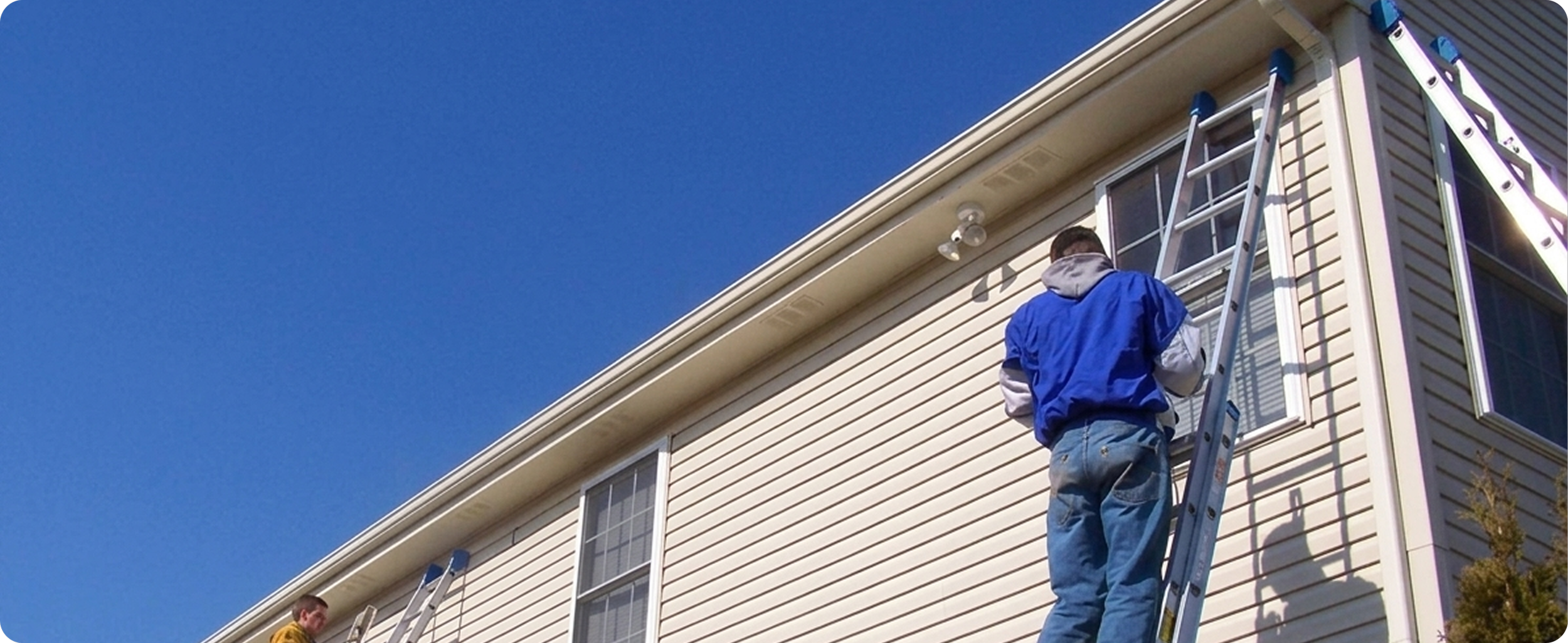 A person in a blue jacket and jeans standing on a ladder, working on the exterior of a beige house with vinyl siding, under a clear blue sky.