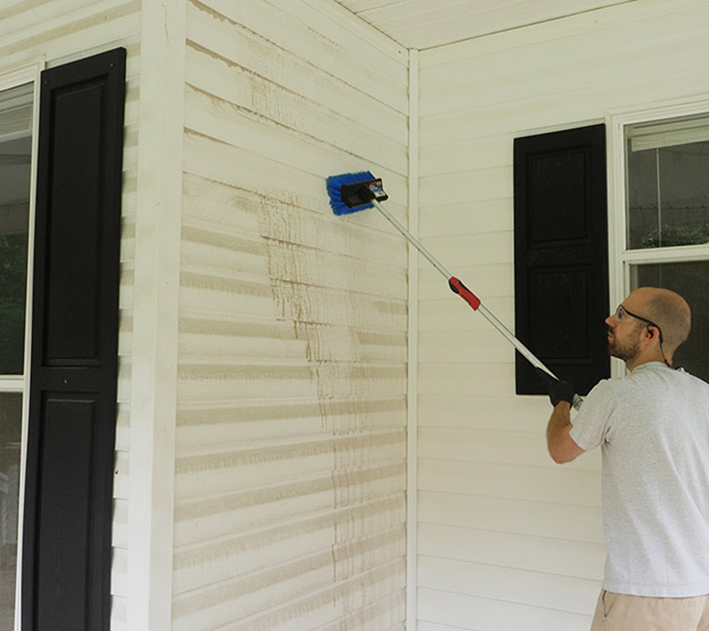 A man wearing safety glasses using a long-handled brush to clean dirty runoff and buildup off white exterior house siding near black window shutters.
