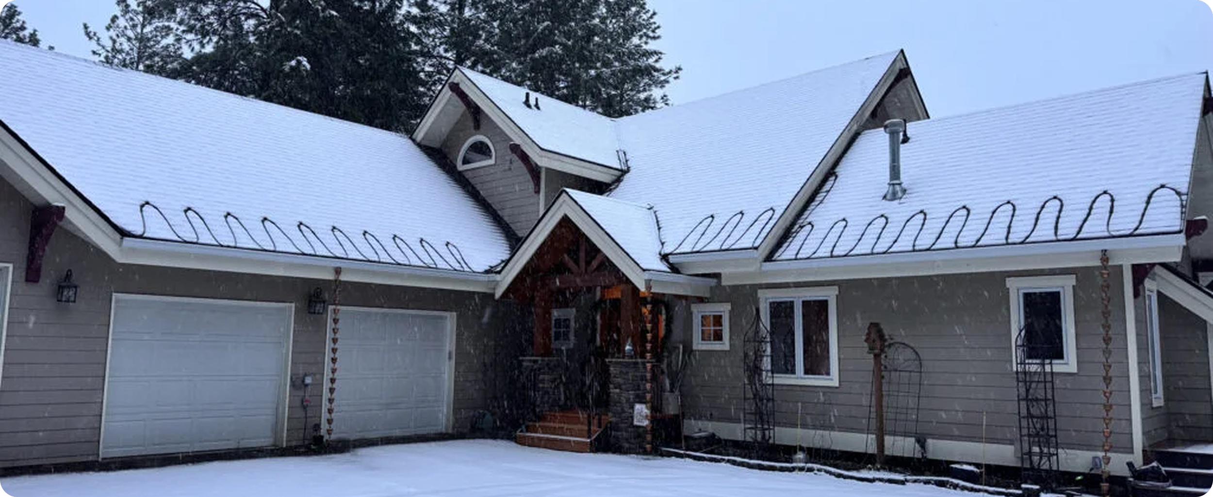 A house with a snow-covered roof, trees in the background, and Christmas lights hanging on the front porch.