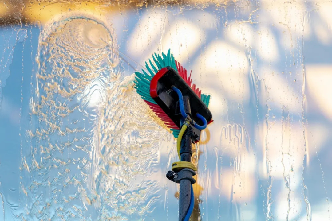 A window cleaner's squeegee with a scrub brush attached, cleaning a glass window with water and soap, creating streaks and bubbles in the glass.