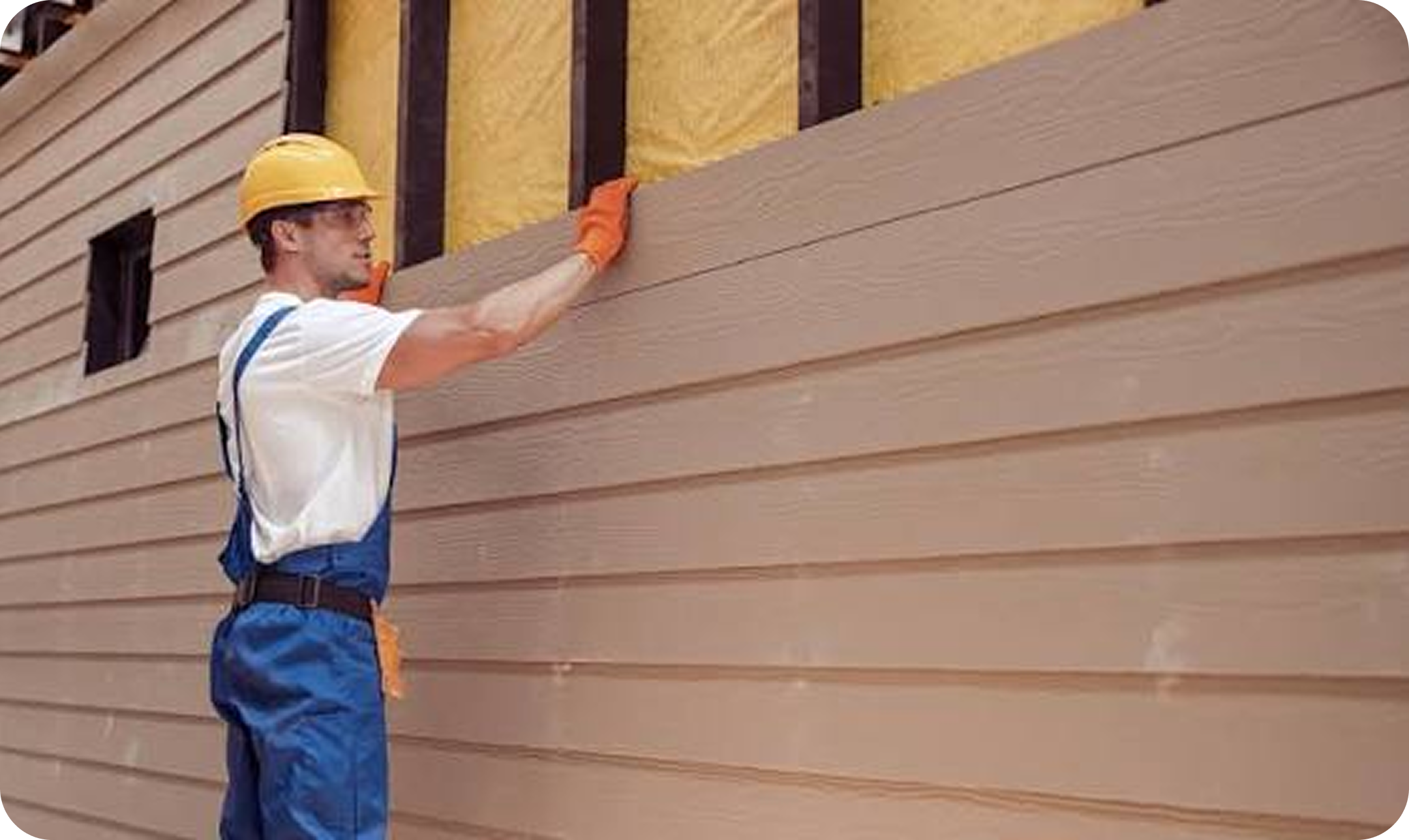 A construction worker wearing a yellow hard hat, safety glasses, an orange glove, a white shirt, and blue work pants is installing siding on the exterior of a house. The siding is a horizontal wooden plank design, and the house wall has some insulation material exposed.
