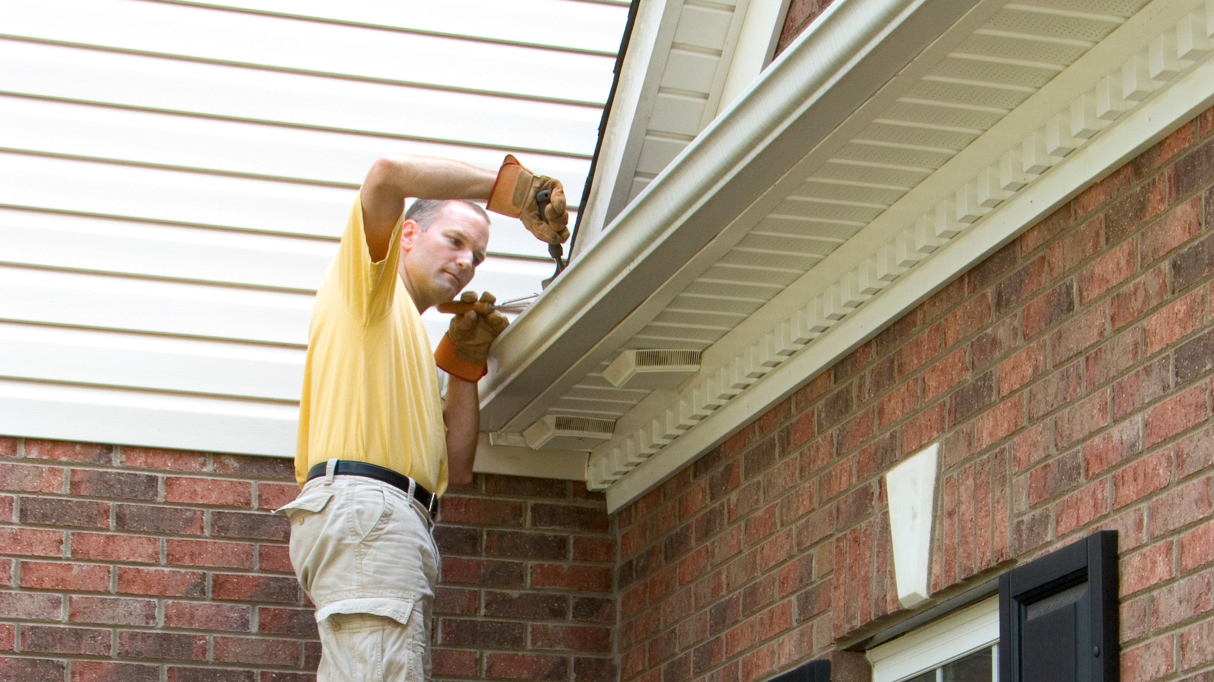 A man wearing a yellow shirt and khaki shorts using a power drill to work on the exterior of a house, standing on a ledge below a white soffit and vent, with red brick walls around him.