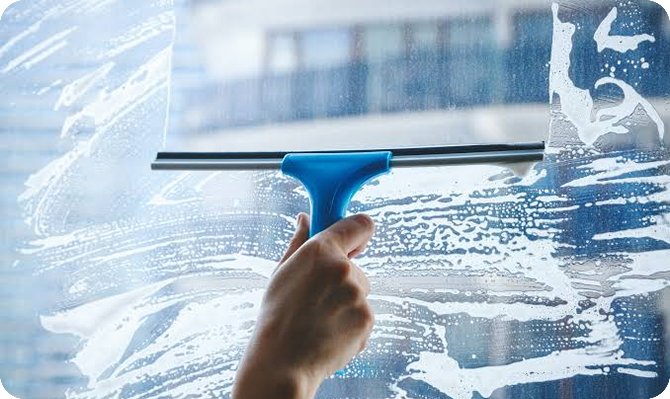 A close-up shot of a hand holding a blue squeegee, dragging it across a foamy glass window to clean the surface.