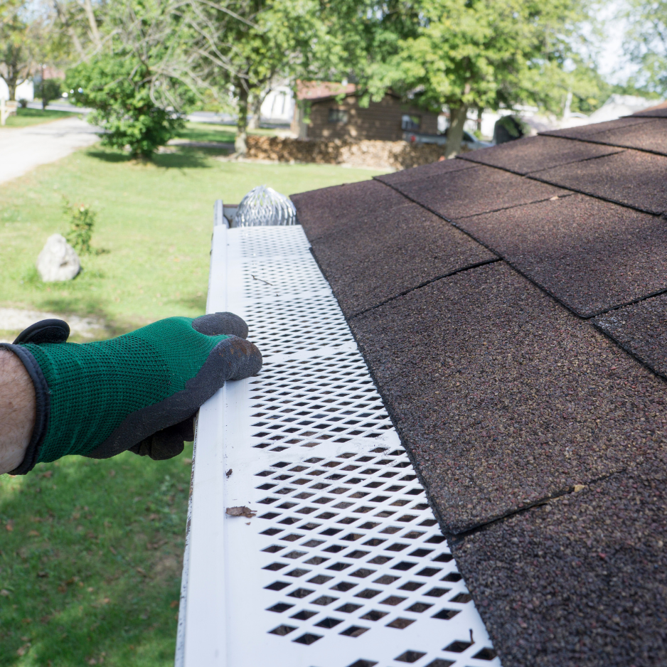 Person wearing green gloves installing or inspecting the white gutter on a house roof with brown shingles, with trees and houses visible in the background.