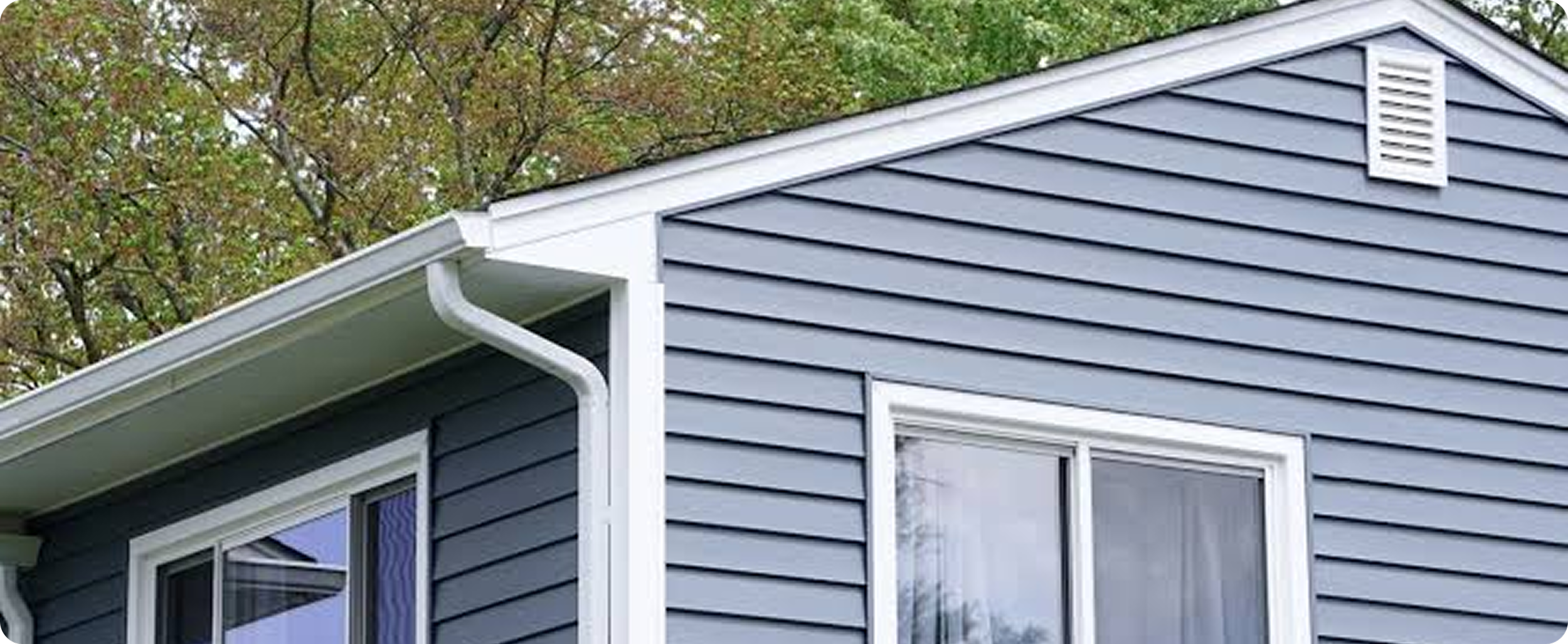 Close-up of the top corner of a blue house with white trim, siding, and window, with trees in the background.