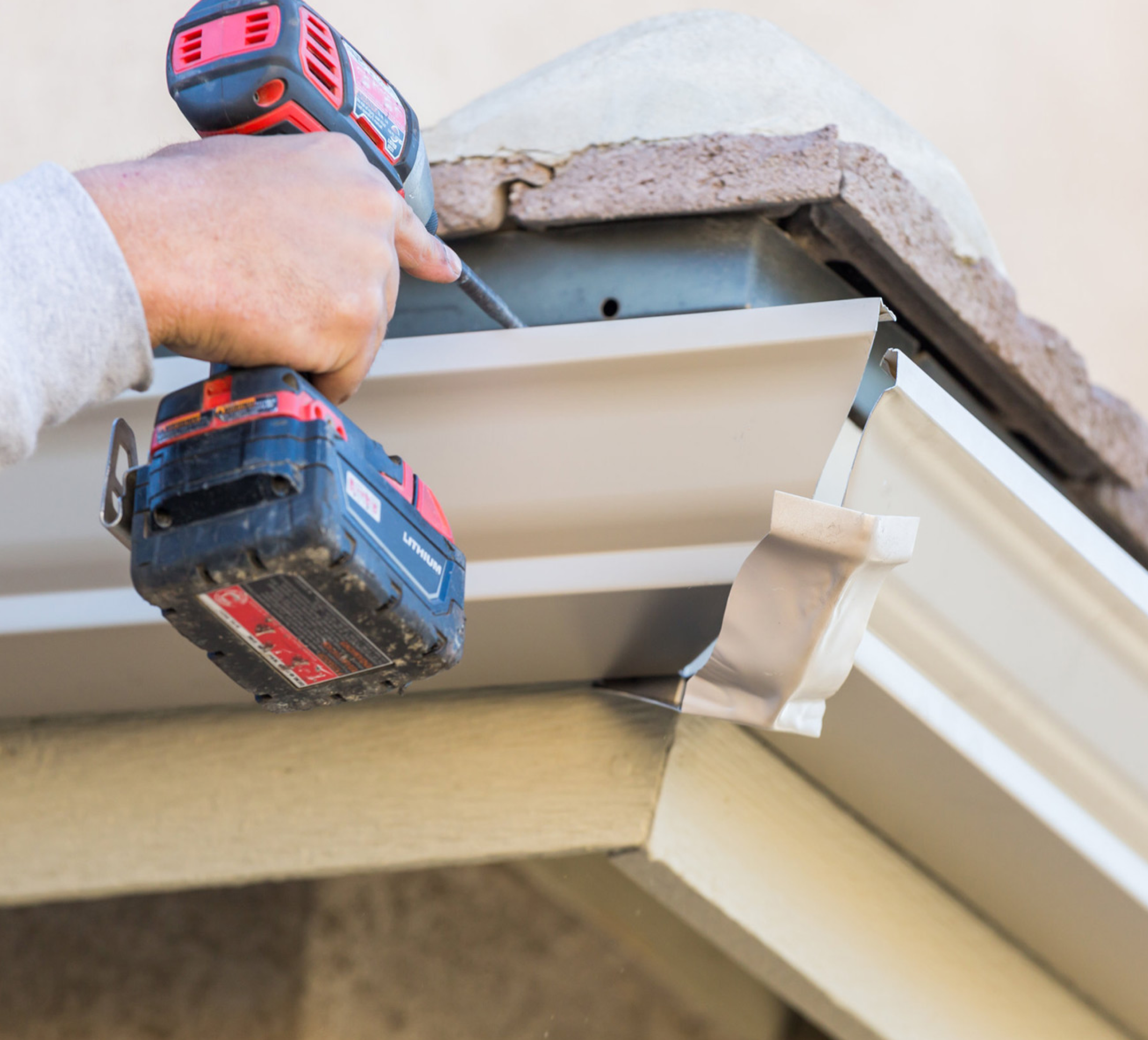 Close-up of a hand holding a red and black cordless drill, drilling into a metal surface, with a beige siding wall in the background.
