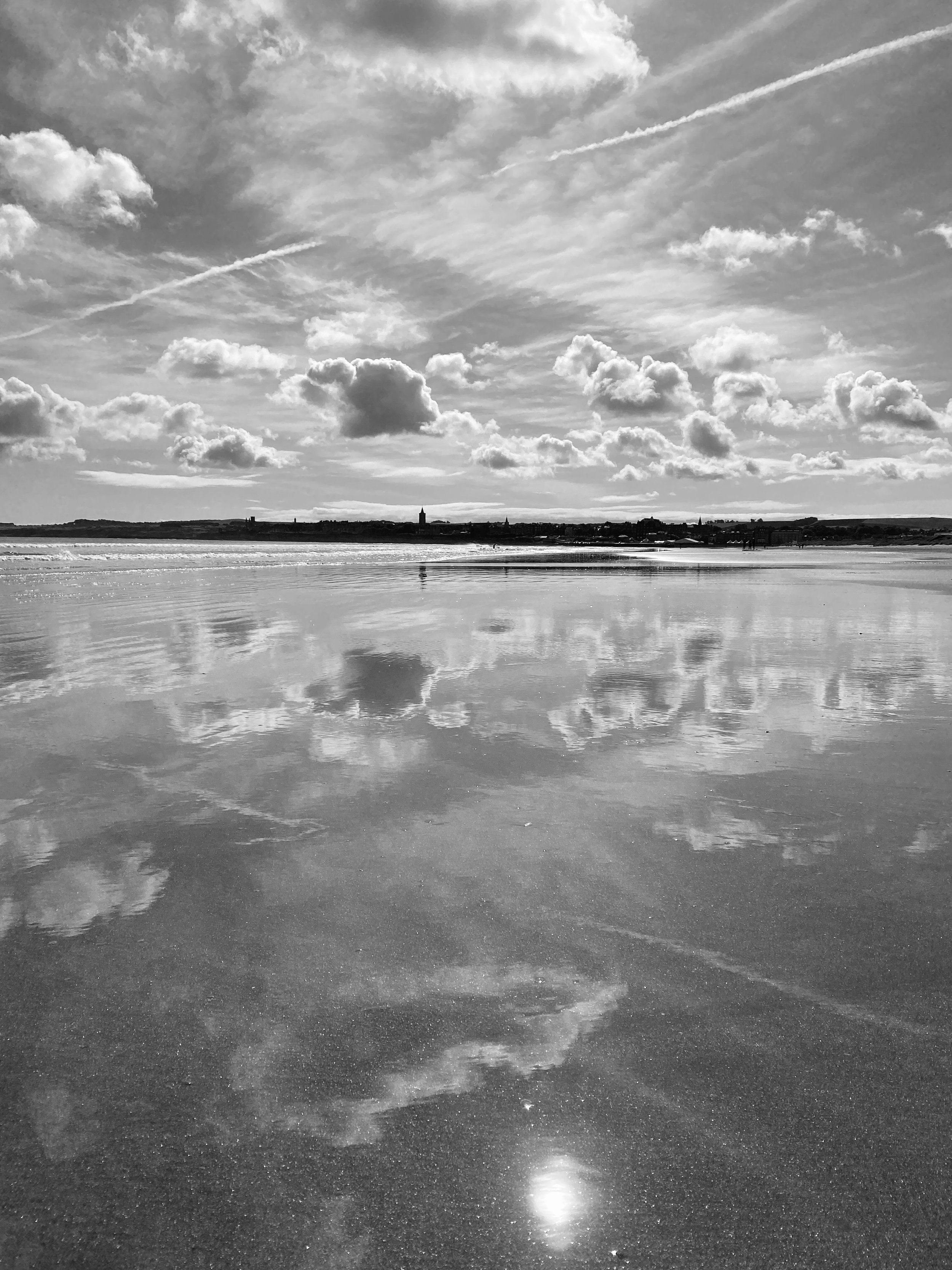 Black and white photo of a beach with reflective water, cloudy sky, and distant shoreline.