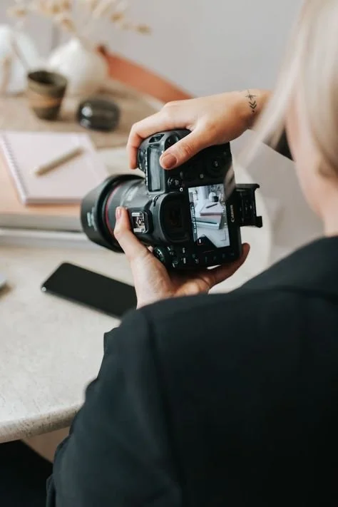 Person holding a professional camera with a flip-out screen, taking a photo of a workspace that includes a notebook, pen, and smartphone on a desk.