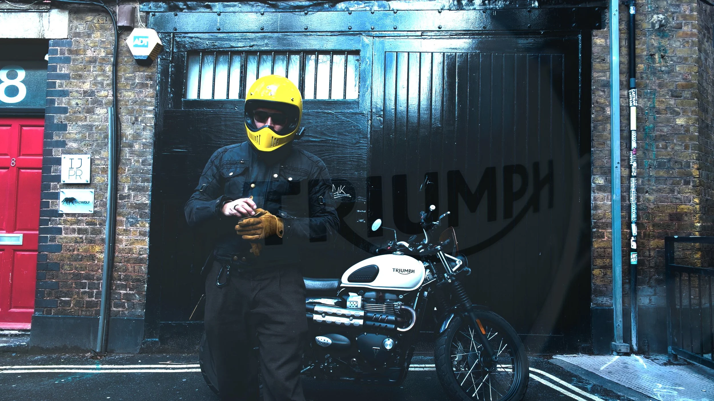 A person in a black jacket, yellow helmet, and sunglasses standing next to a white Triumph motorcycle in front of a black garage door with a faint Illy logo, on a city street with brick walls and a red door.