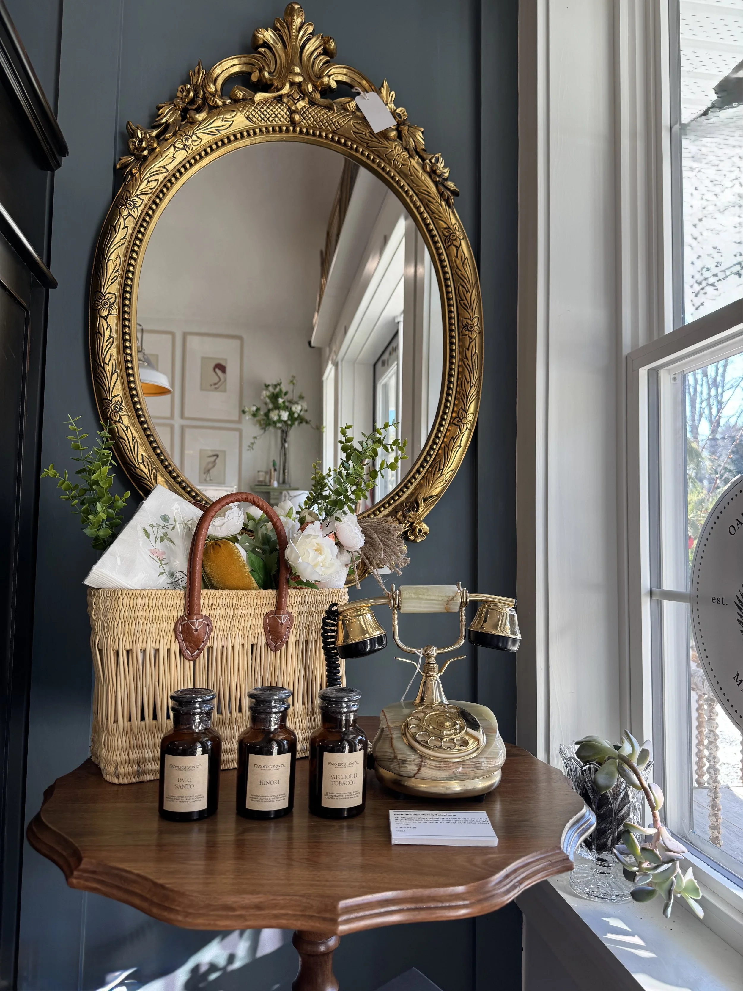 A decorative vintage mirror with ornate gold frame, a wooden table with three amber glass bottles labeled Palo Santo, Hinoki, and Patchouli Tobacco, a small tray with candles, a vintage telephone, and a wicker basket filled with flowers and greenery. A window with natural light and a small potted succulent are visible in the background.