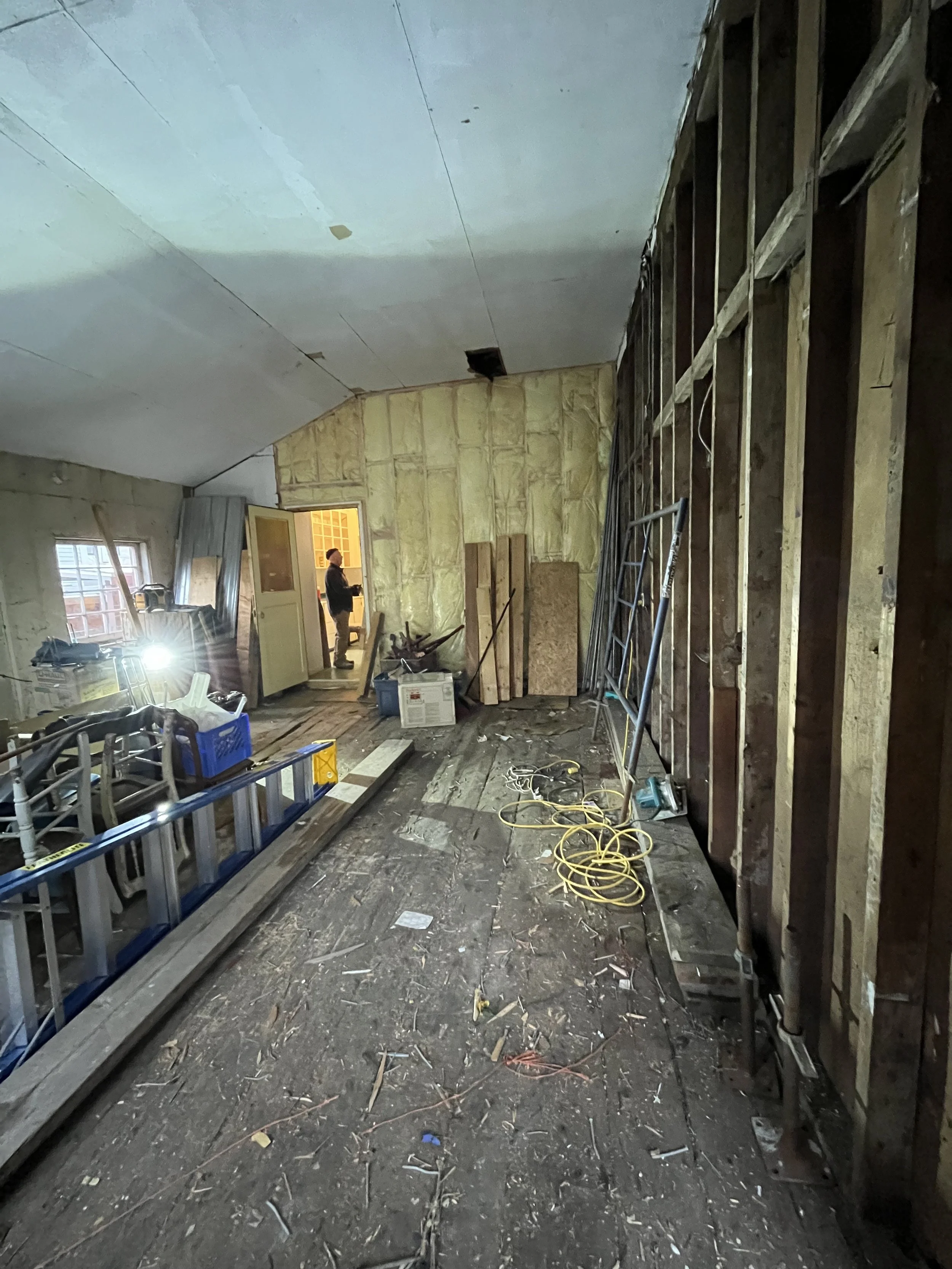 The room is under construction with wooden framing on one wall, construction tools, cables, and debris on the floor, a ladder leaning against the wall, and a person standing near an open doorway in the background.