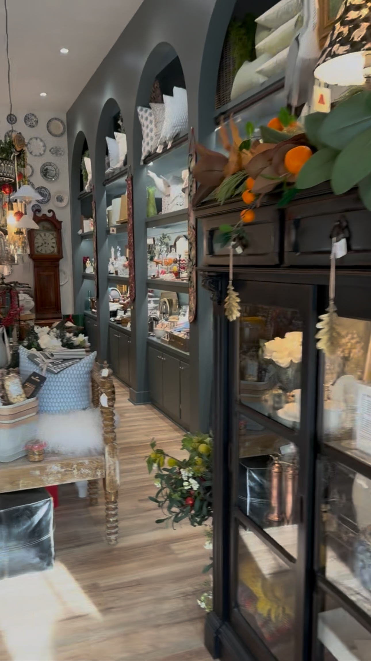 Interior of a vintage-style boutique with display shelving, decorative plates on the wall, a tall grandfather clock, and various home decor items like pillows, lamps, and flowers.