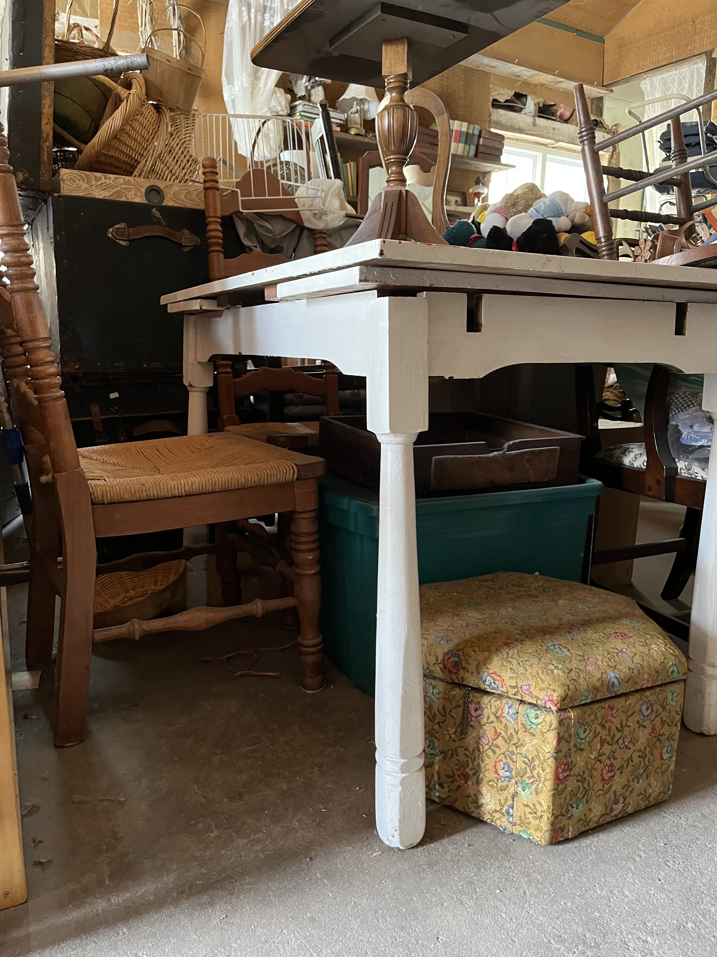 Surplus furniture and household items in a cluttered storage or garage space, including wooden chairs, a small tiled table, a upholstered ottoman, and miscellaneous household objects.