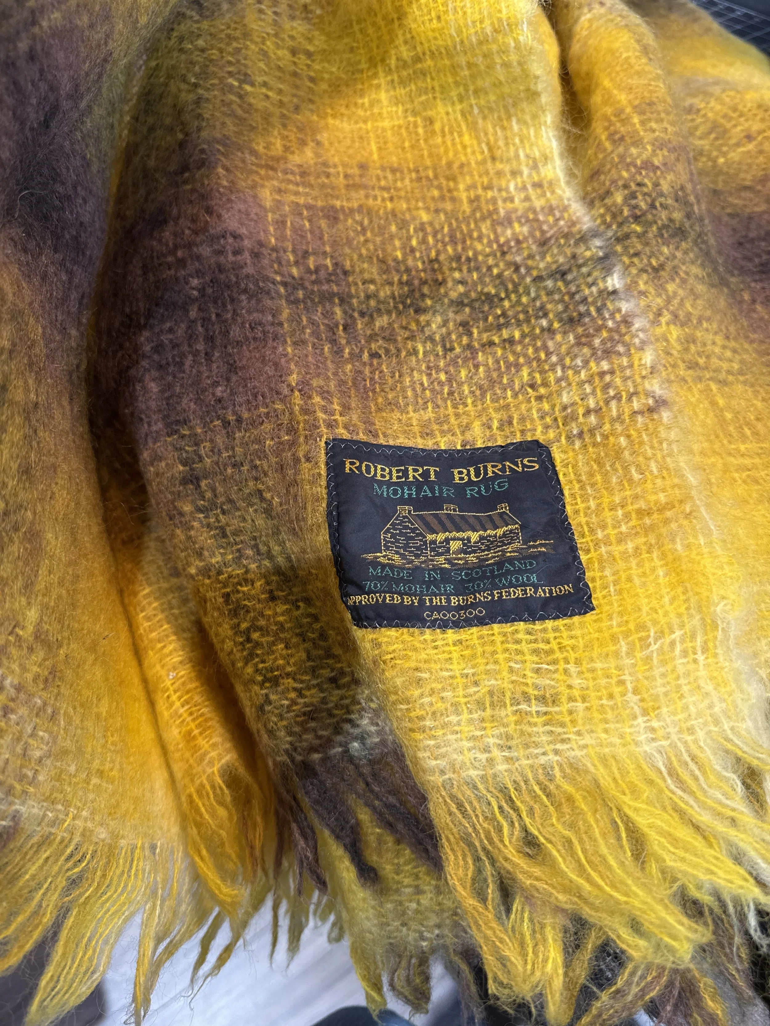 Close-up of a yellow, brown, gray, and purple mohair wool scarf with a label that reads 'Robert Burns Mohair Rug Made in Scotland 70% Mohair 30% Wool'.