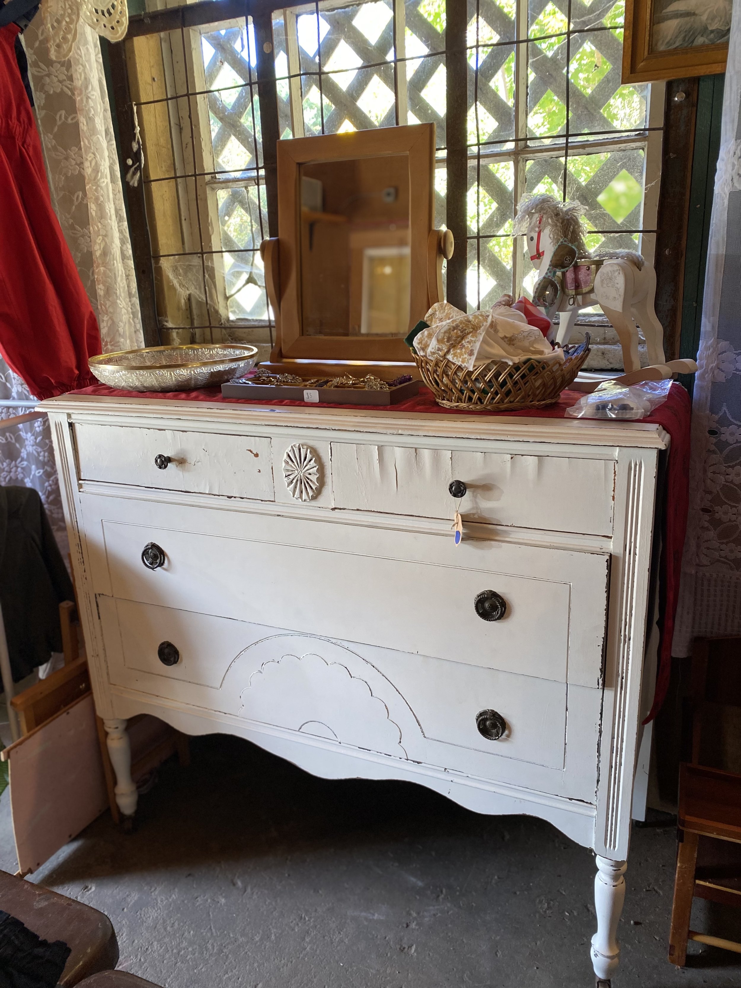 A white wooden dresser with four drawers and decorative carvings, topped with various items including a small wooden mirror, jewelry, a basket with cloth, and a porcelain horse toy, situated in front of a window with lattice-patterned bars and lace curtains.