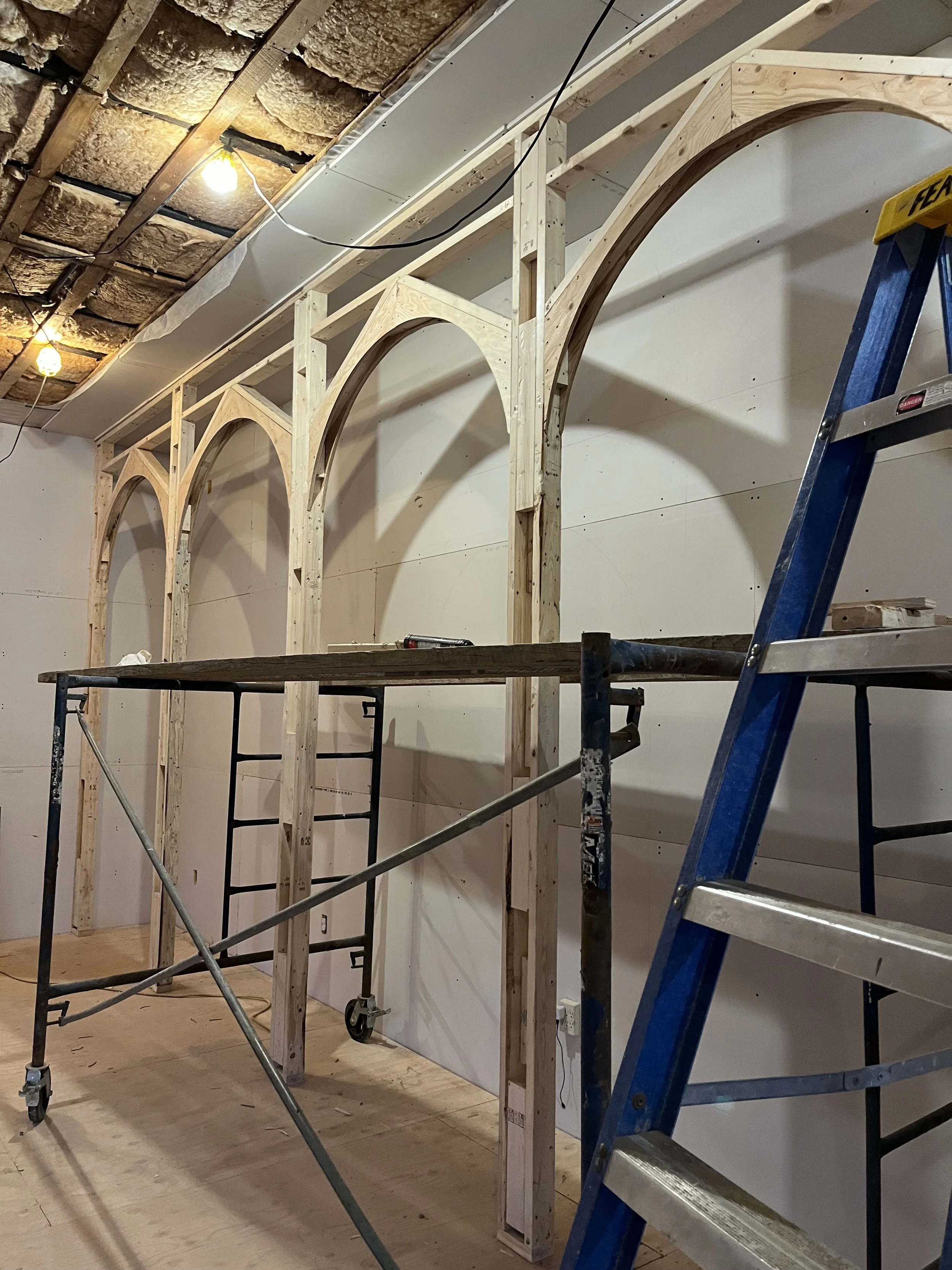Wooden construction frame with arches being built on a wall, scaffolding and ladders present, ceiling exposed with insulation and construction lighting.