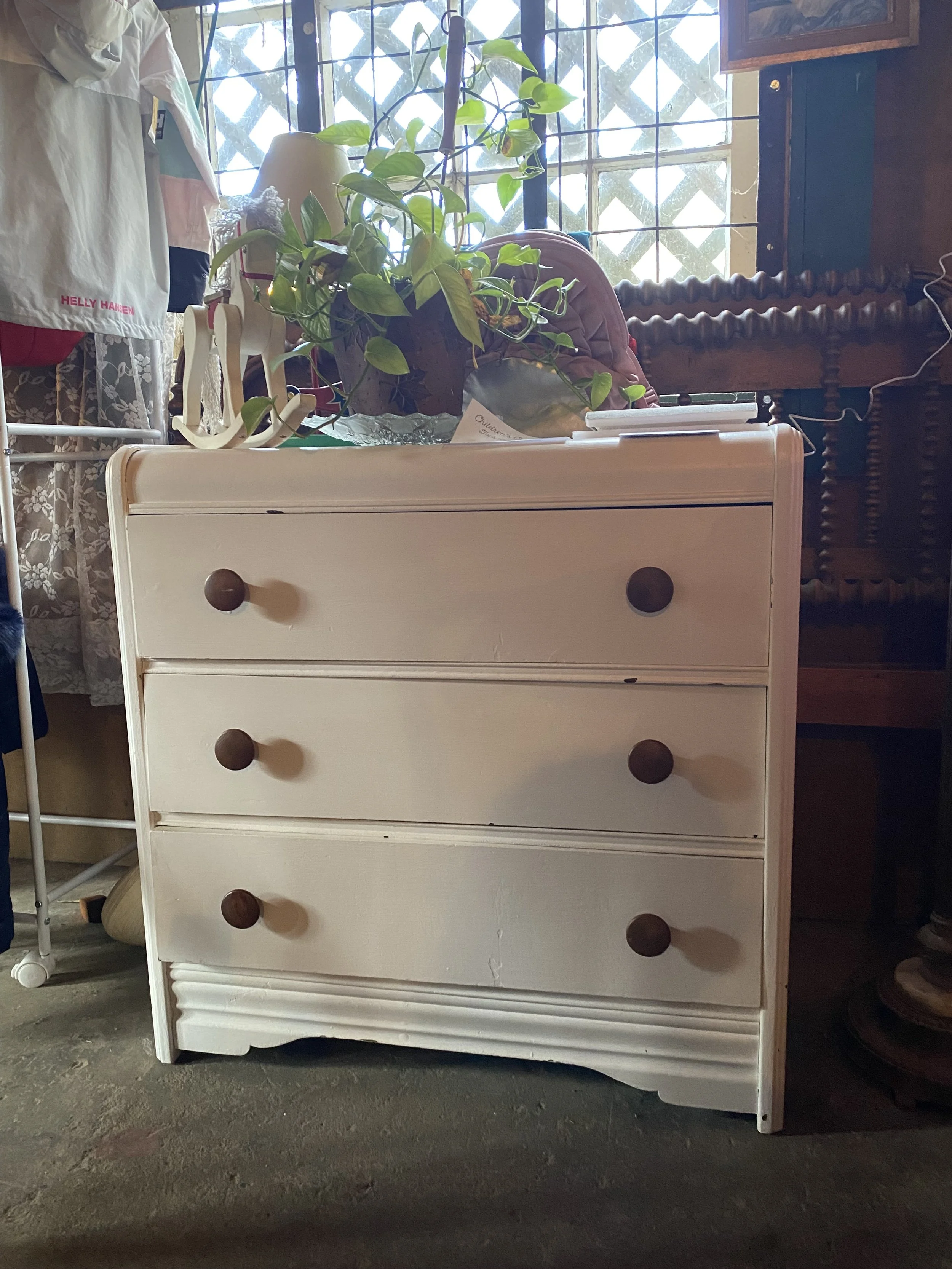 A white wooden dresser with four drawers and round dark knobs, outdoors on a concrete surface. A potted green plant on top, with a pink object behind it, in front of a window with metal bars and a lattice pattern. Items like a lamp, folded clothes, and a sign are also visible on or near the dresser.
