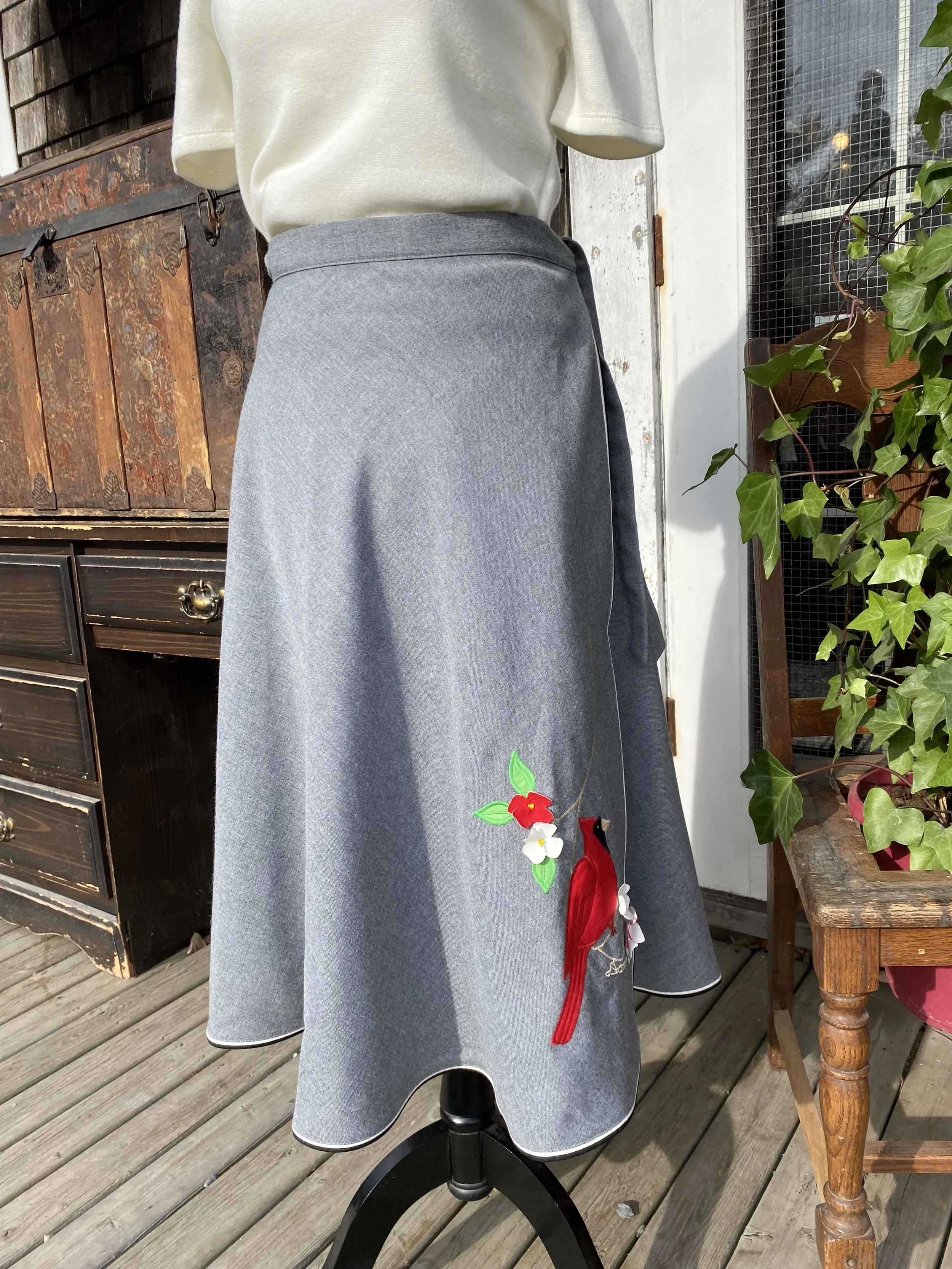 A mannequin dressed in a gray skirt with floral embroidery and a cream-colored top, displayed outside on a wooden deck, with potted plants and wooden furniture in the background.